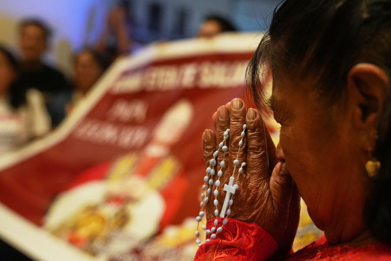 A woman prays with rosary beeds in front of a banner of Pope Leo XIV