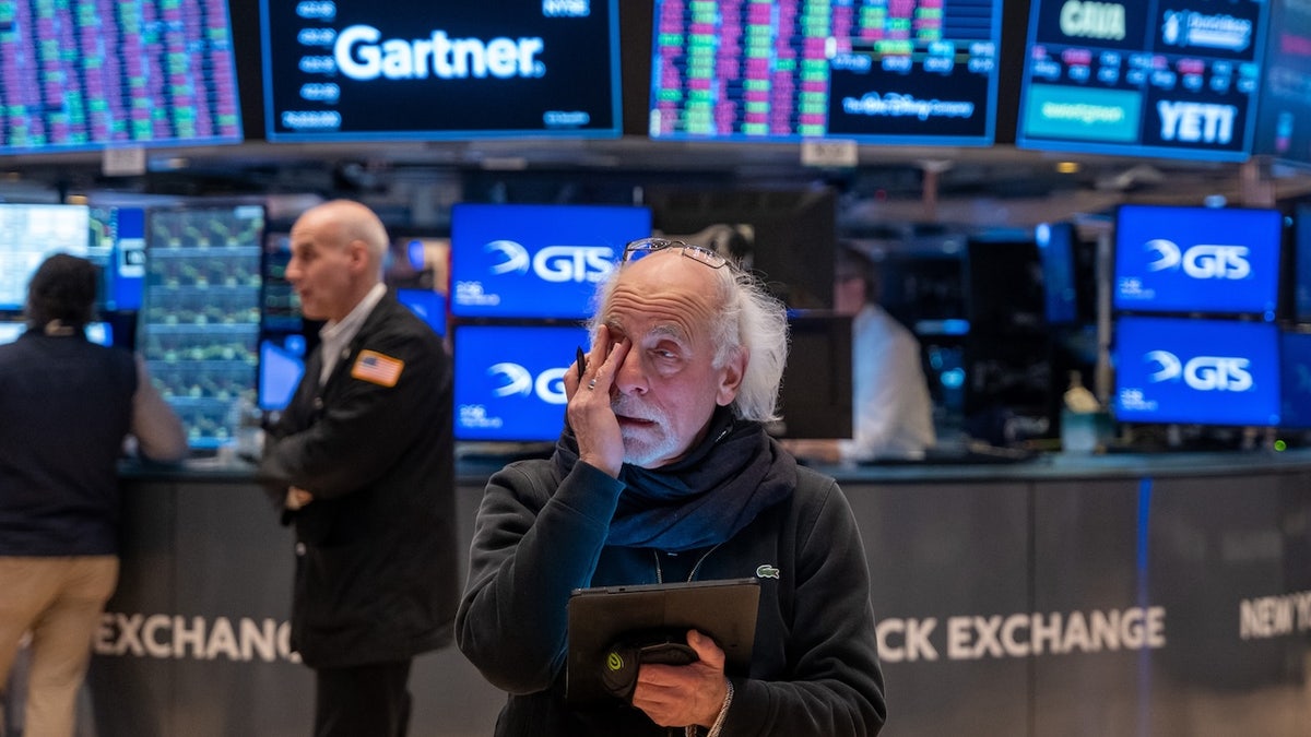 A visibly concerned trader holds a tablet and rubs his eye on the floor of the New York Stock Exchange, with colorful stock tickers and monitors flashing behind him.