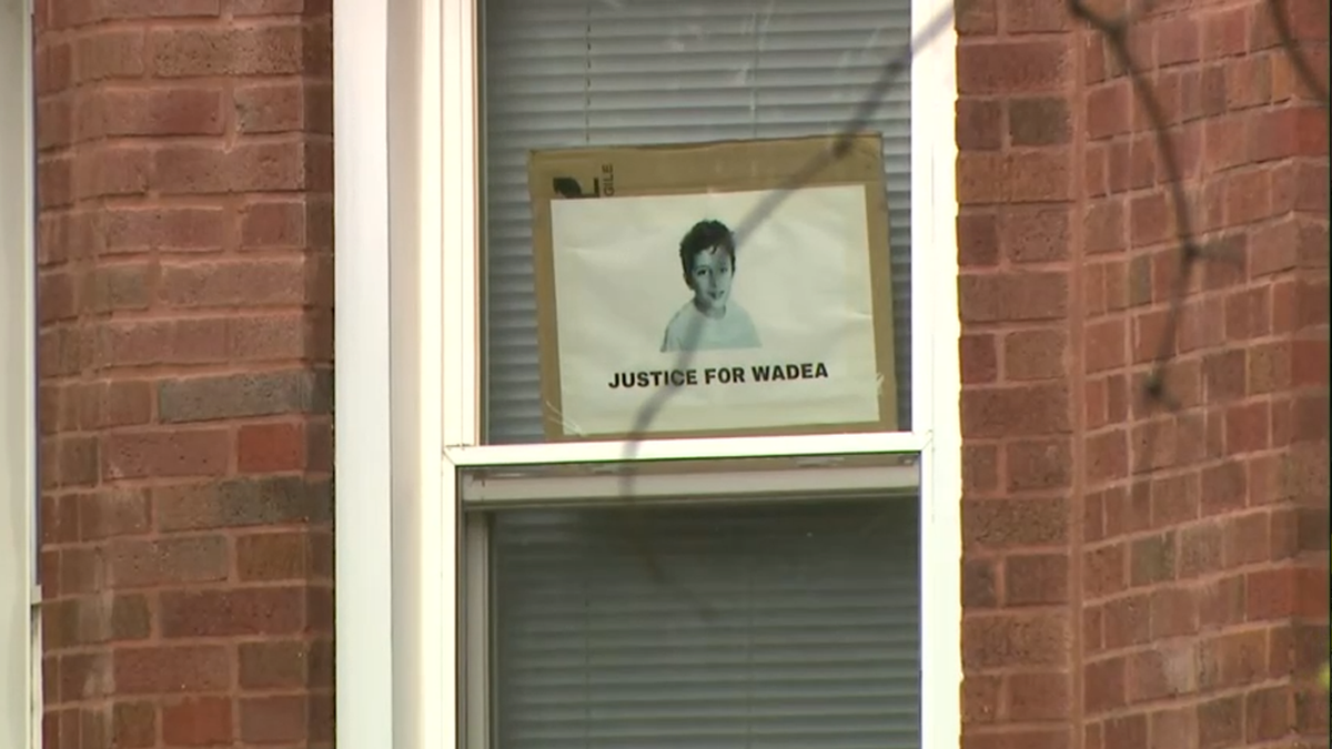 A black and white photo of a little boy with the caption "Justice for Wadea" is propped up against a window with the blinds closed behind it, set in a brick wall.