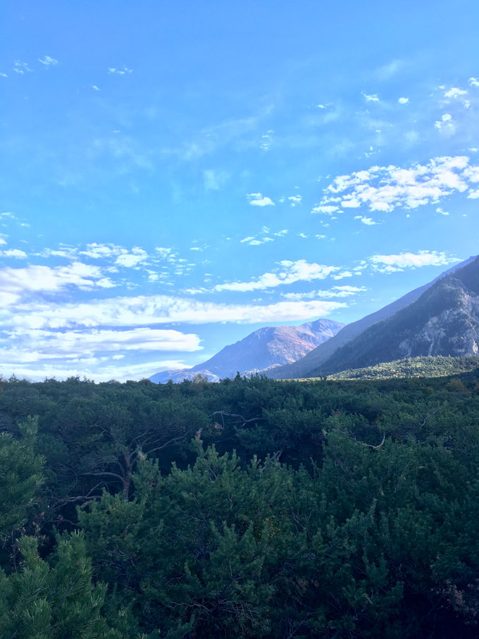 A mountain landscape with layered hills receding into the distance under a blue sky with scattered white clouds and a foreground filled with dense green vegetation