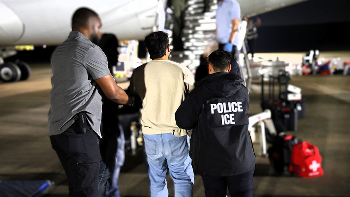 ICE agents escort a male detainee in hand restraints up a stairway to a deportation plane