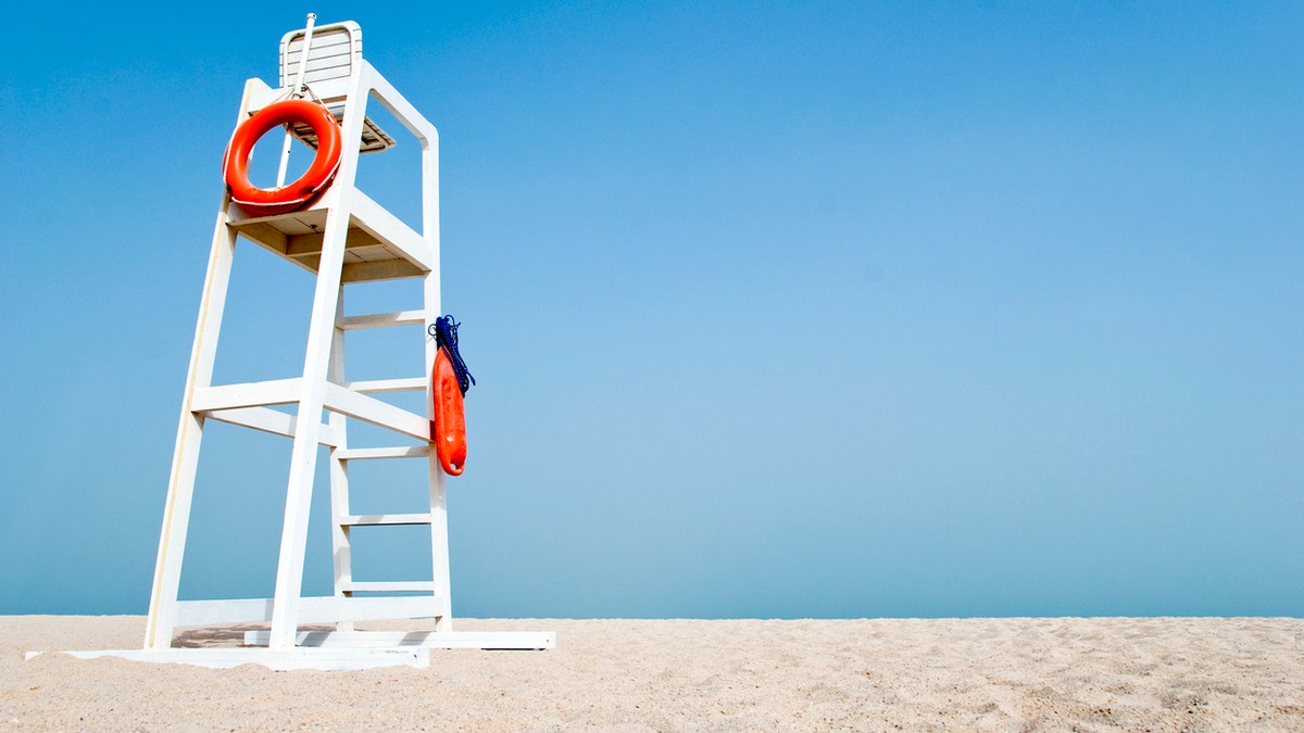 Empty white lifeguard chair on an empty beach with orange life buoys hanging on the side.The chair stands against a clear blue sky in the sand.