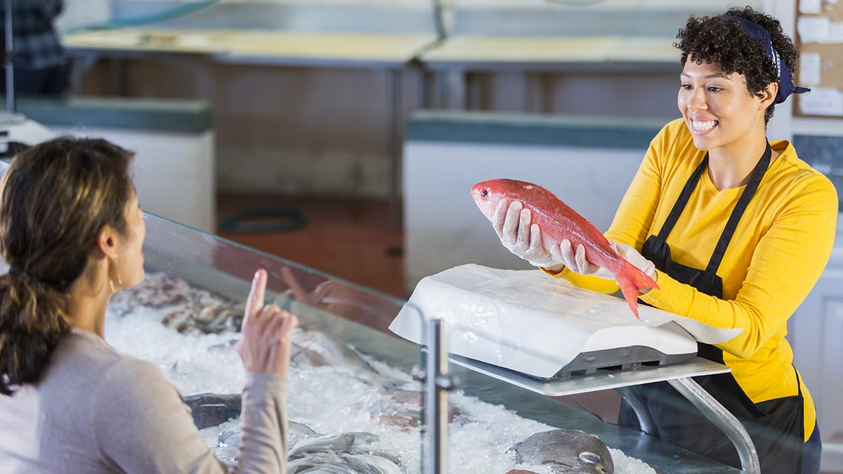 Woman in seafood market helping customer, woman shopping asking question seen from behind as worker holds out big pink fish