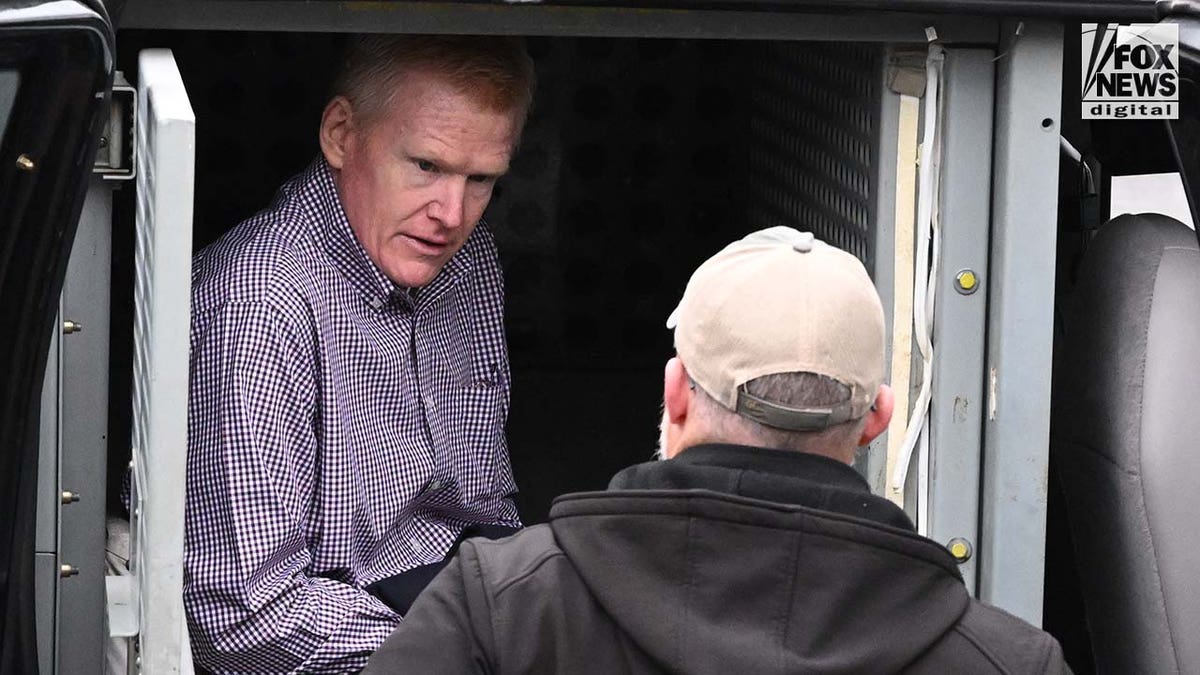 A man wearing a plaid shirt exits a police vehicle surrounded by officers.
