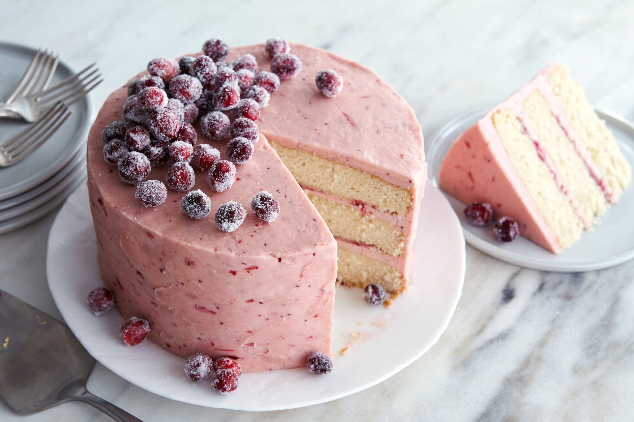 A pink layer cake topped with sugared cranberries sits on a white plate. To the top left are a stack of plates topped with forks and a cake server. To the right is a plate holding a slice of the same cake.