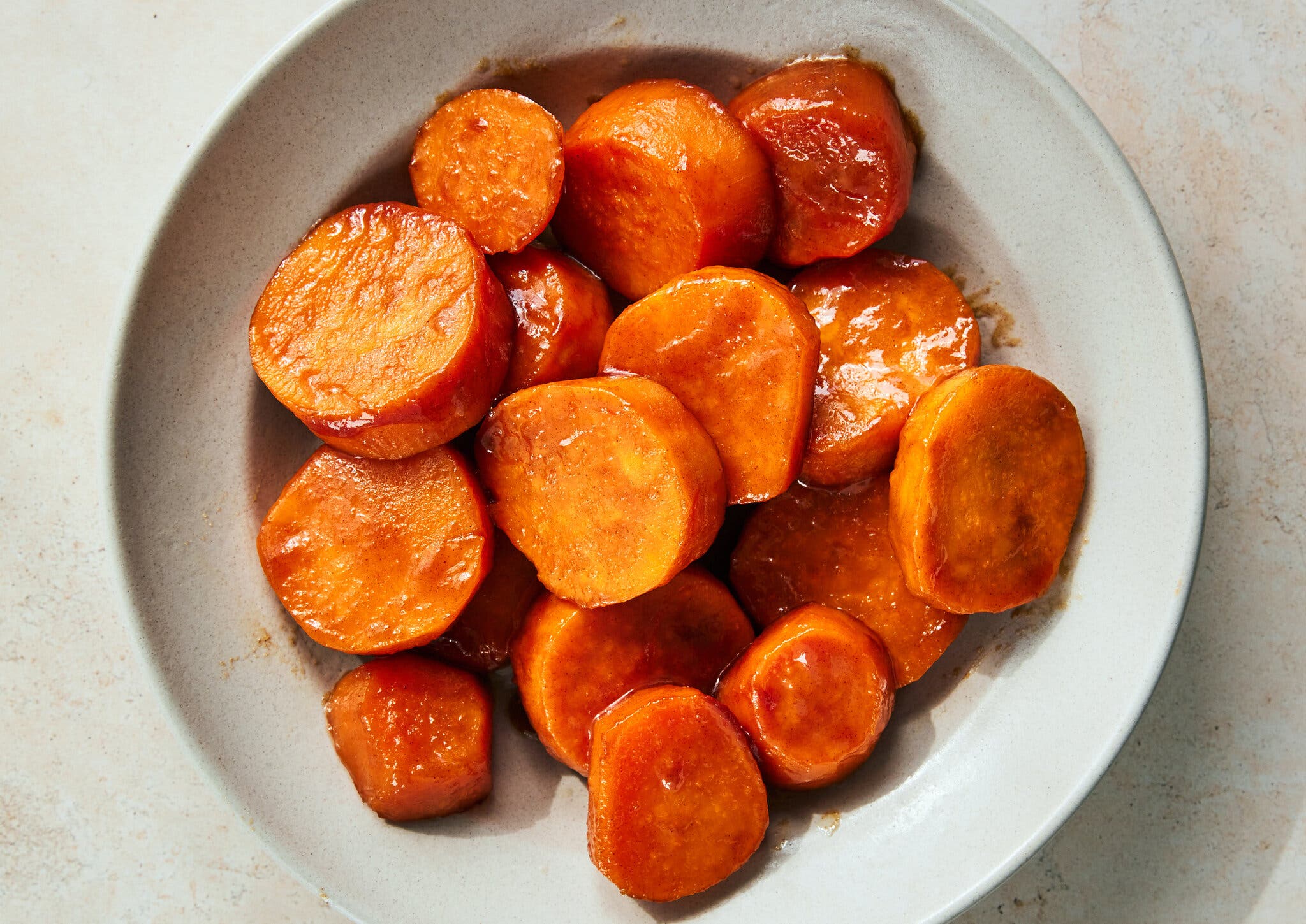A white plate sitting against a light marbled background holds a number of round sweet potato slices coated in a glaze.
