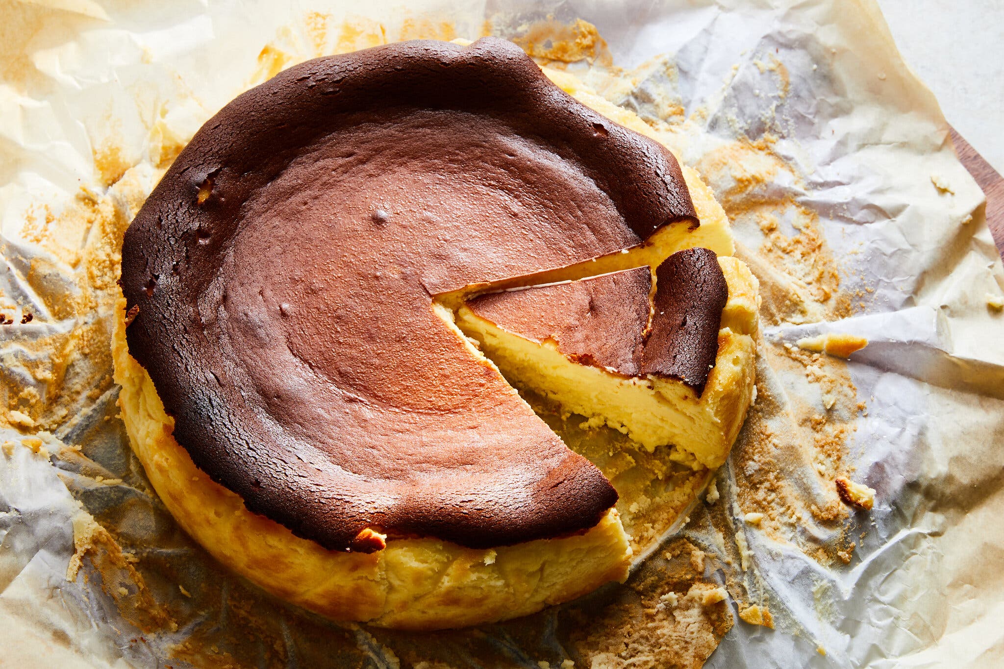 A Basque cheesecake with a brown, burnished crust on parchment paper. There is a slice cut from the cheesecake.