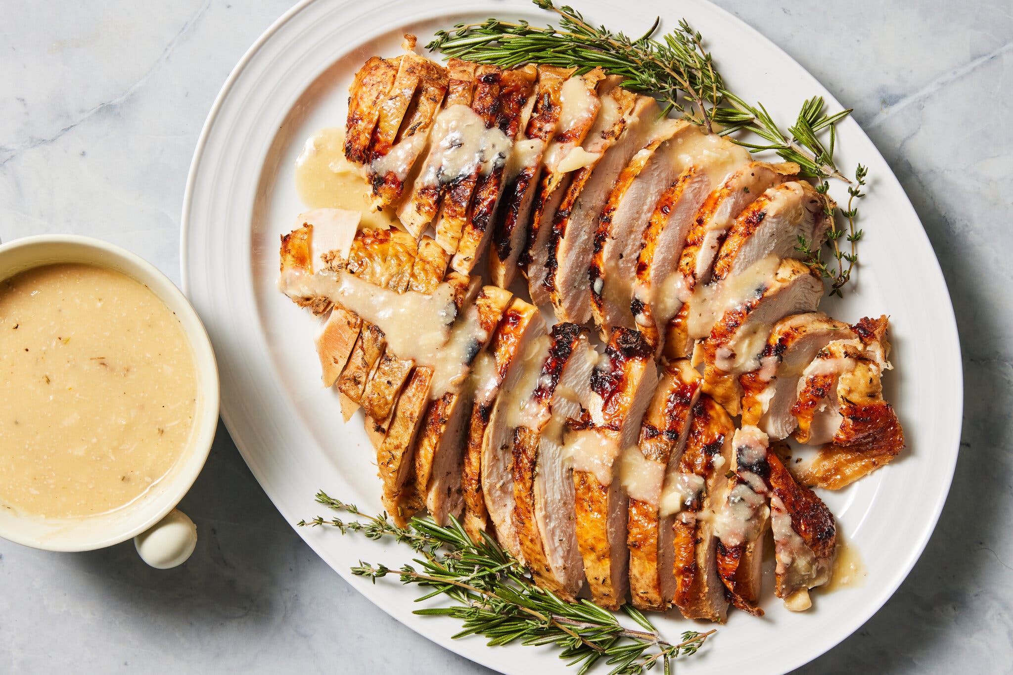 An overhead image of a plate of sliced turkey covered in gravy, with rosemary sprigs as garnish. There is a dish of gravy to the left.