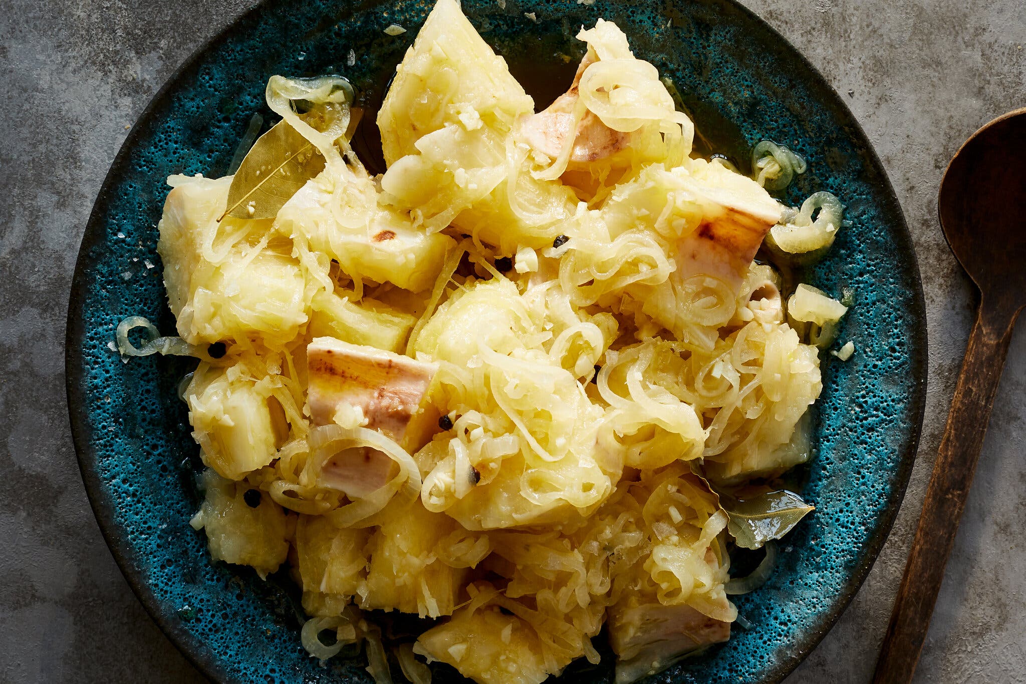An overhead image of a blue and black plate filled with yucca dressed with mojo, a sauce made with onion, garlic, peppercorn bay leaves and vinegar. A worn wooden spoon sits to the right hand side of the plate.