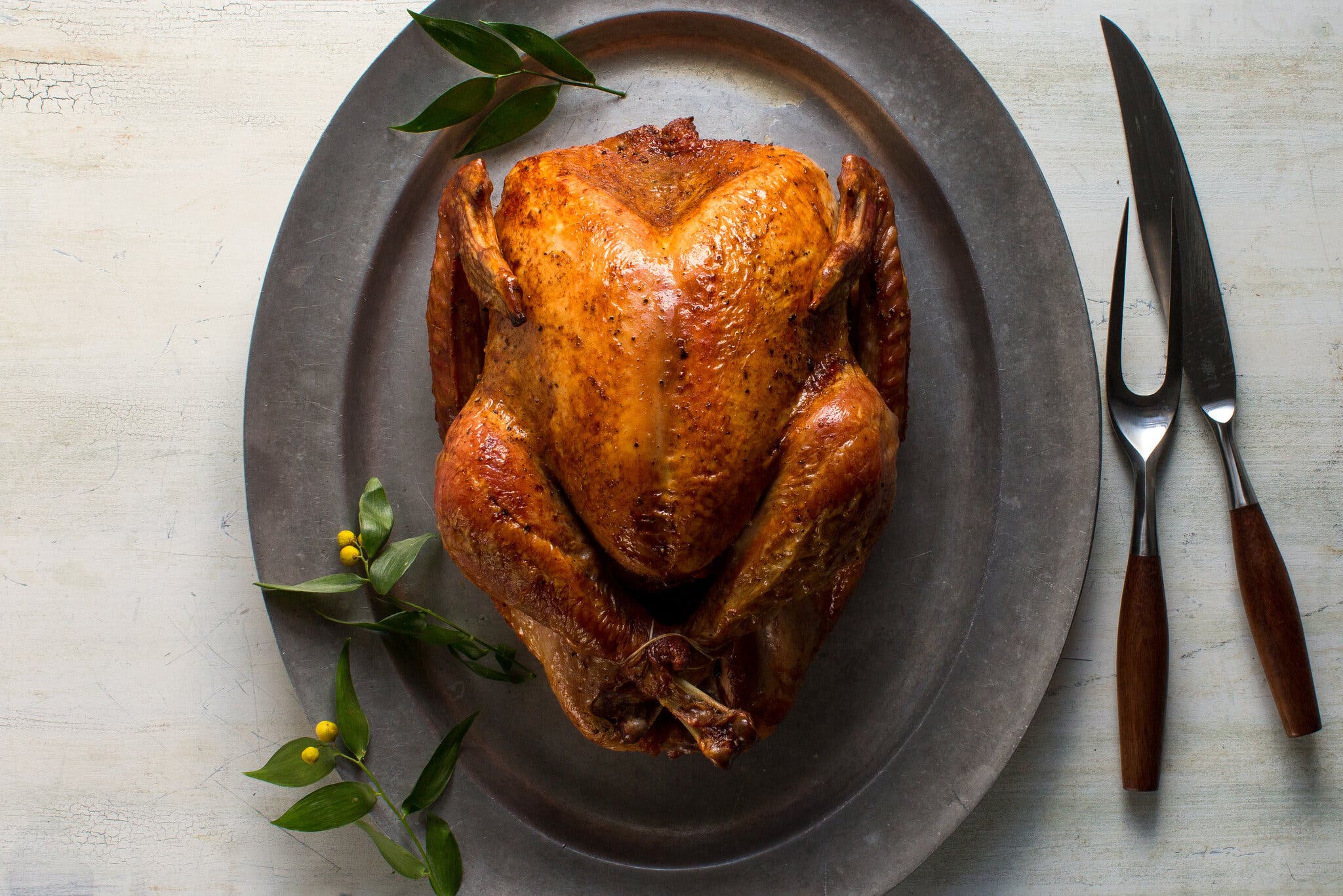 An overhead image of a roasted turkey on a gray platter. A wood handled carving set sits to the right. Leafy green garnishes dot the left hand side.