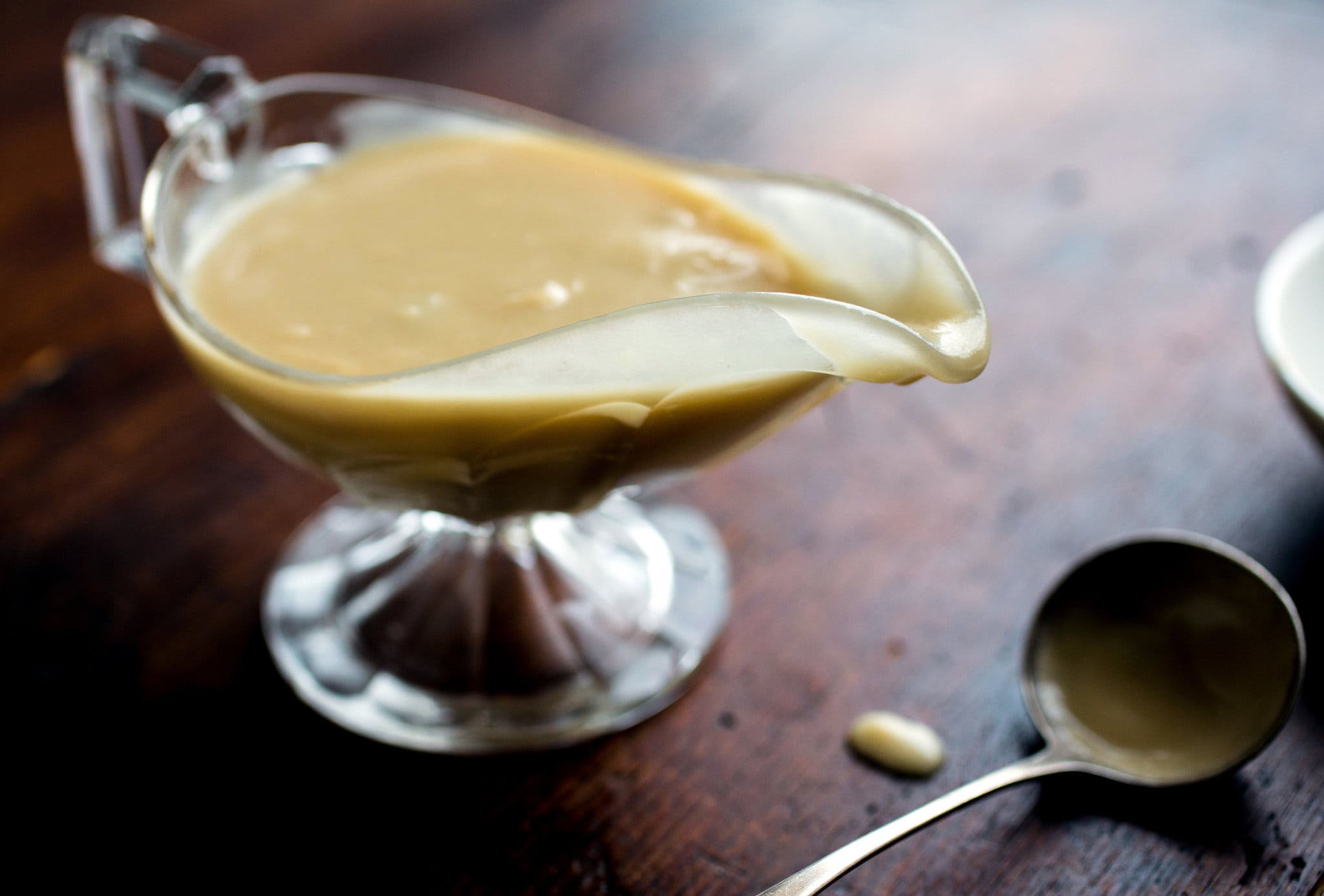 A clear gravy boat sitting on a wooden countertop holds a tan gravy. To the bottom right corner of the frame is a serving utensil coated in gravy.