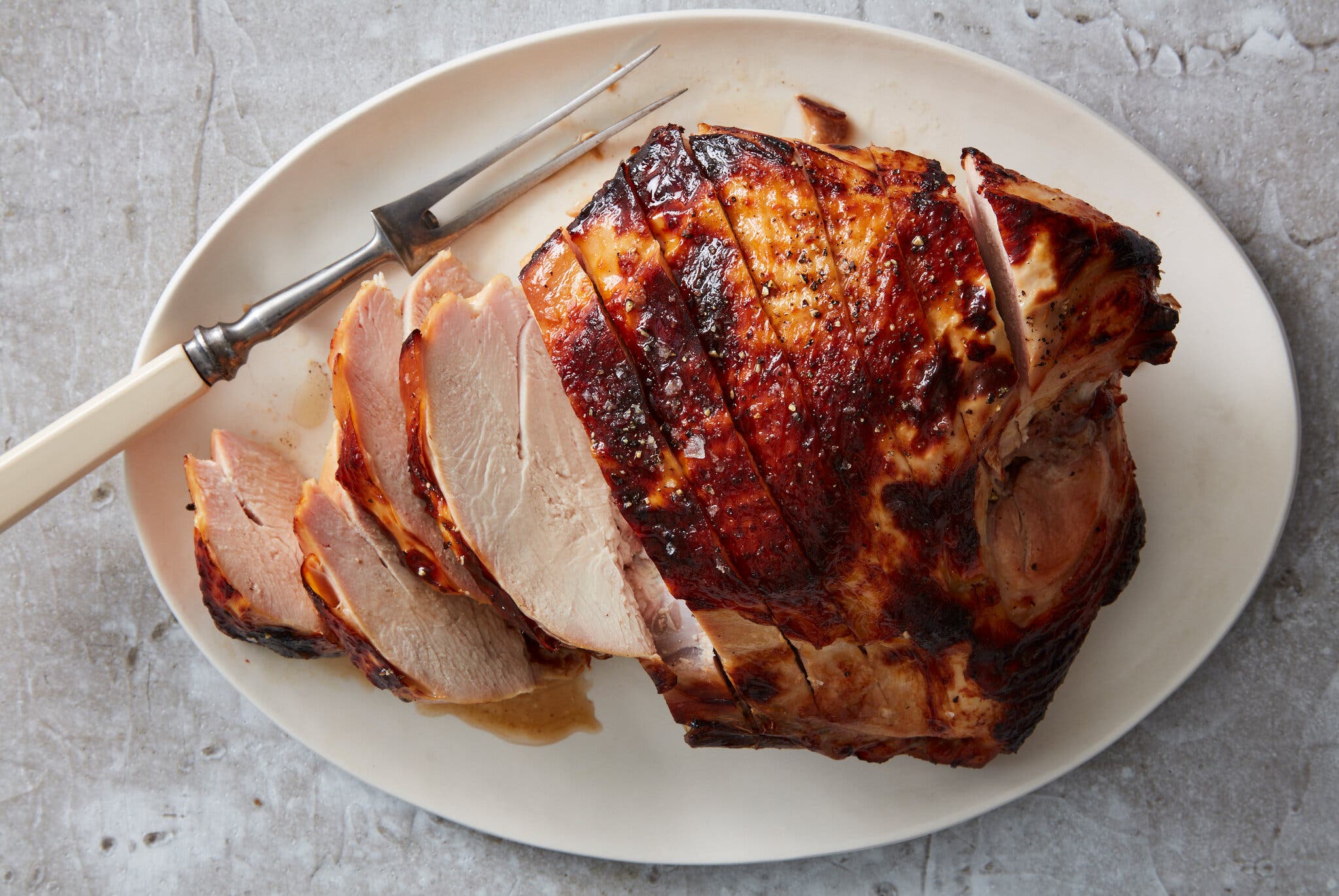 An overhead image of a white oval platter on a gray marble background topped with a burnished turkey breast that is partially sliced. A two-pronged serving fork is balanced on the edge of the plate.