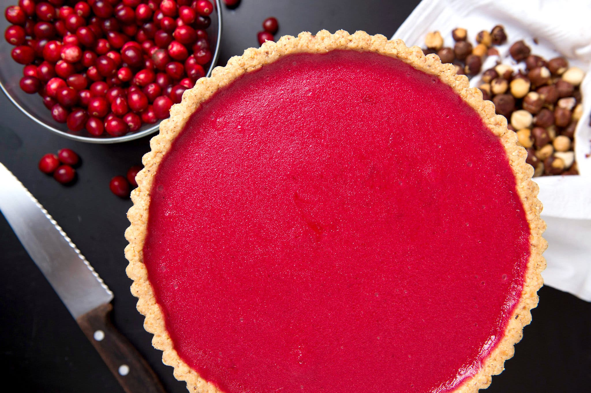 An overhead view of a bright red cranberry tart. A bowl of cranberries and some hazelnuts sit just above it on a table.