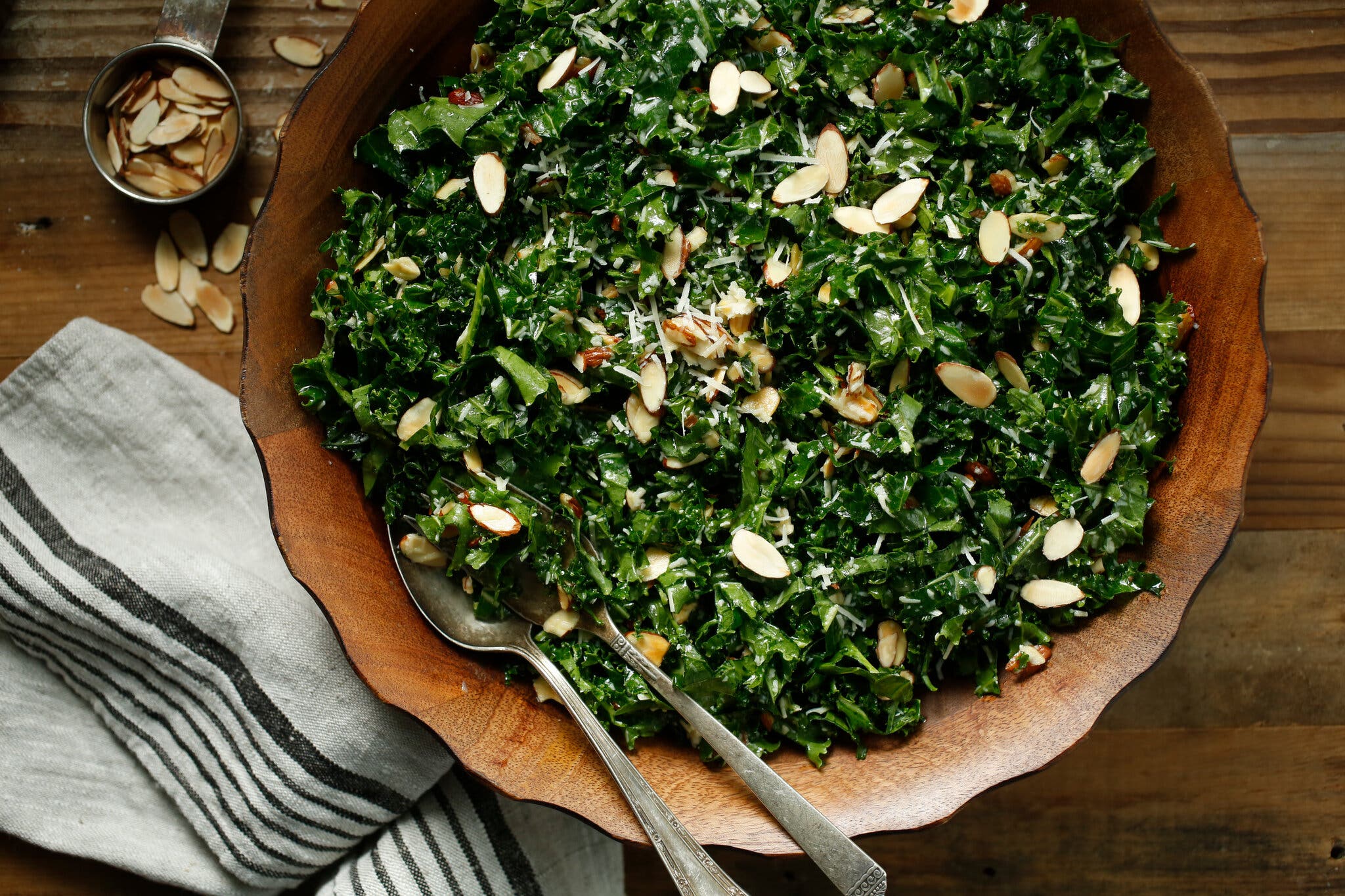 A wooden serving bowl holds lemon-garlic kale salad with slivered almonds and Parmesan. A silver spoon and fork are in the bowl. Additional almonds are in a small measuring cup nearby, as is a striped napkin.