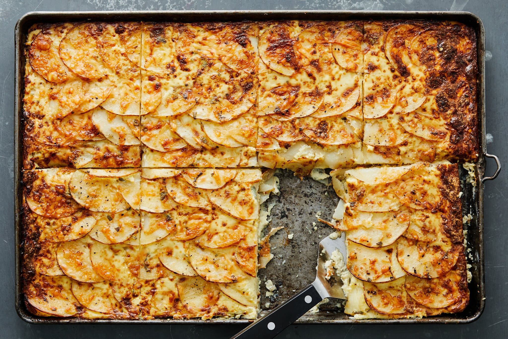An overhead image of a sheet-pan of golden brown, scalloped gratin. The gratin has been cut into eight pieces, and one has been removed.