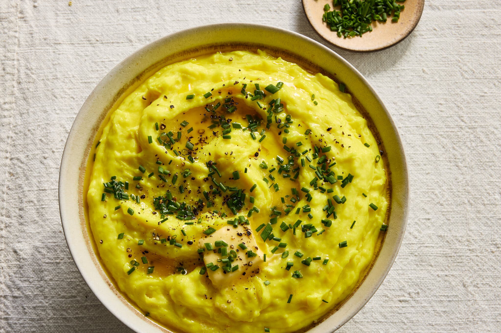 A close-up, overhead view of bright yellow mashed potatoes topped with melted butter, fresh chives and black pepper.