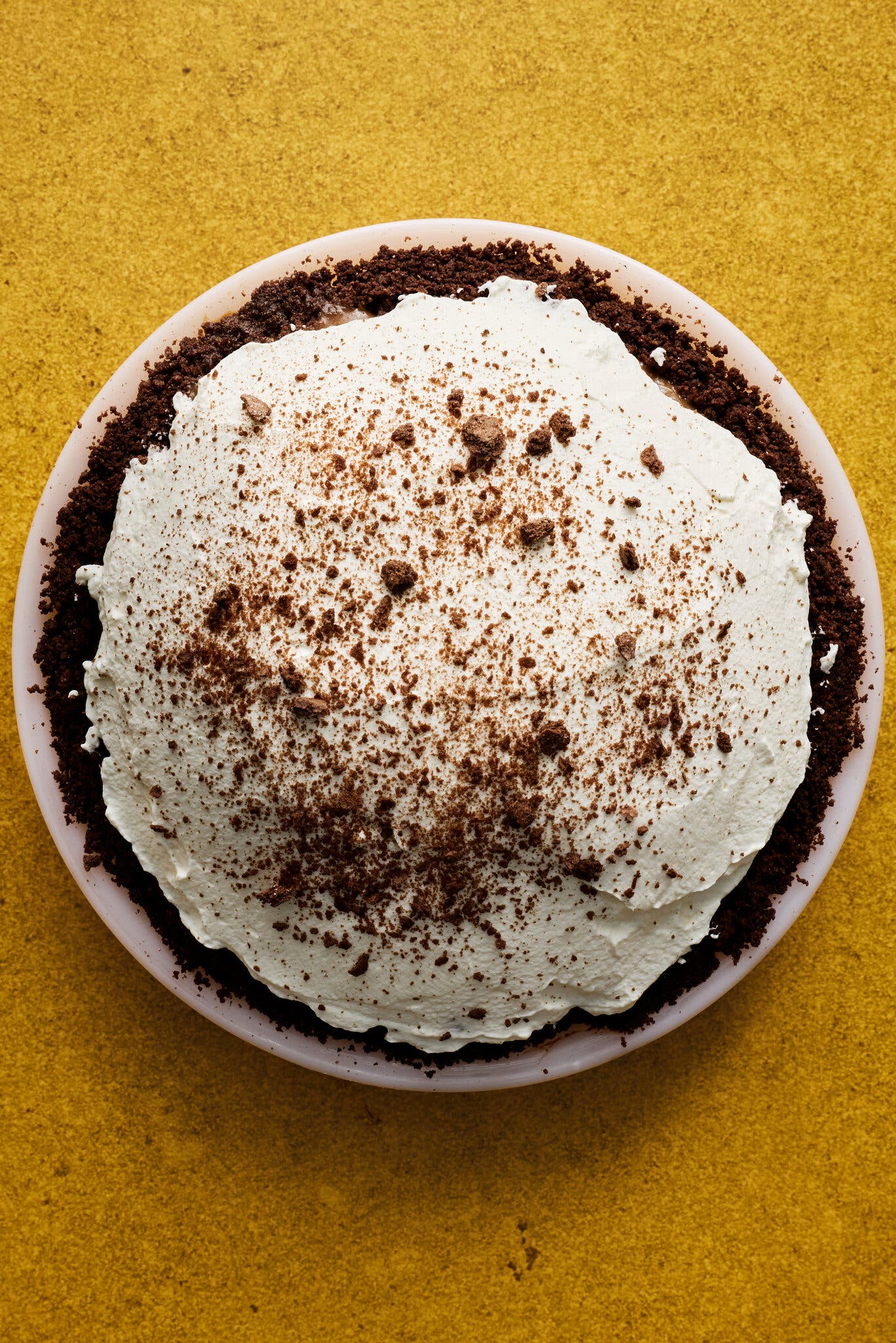 An overhead shot of a pie with a dark, crumbly crust and a mountain of whipped cream topping against a yellow background.