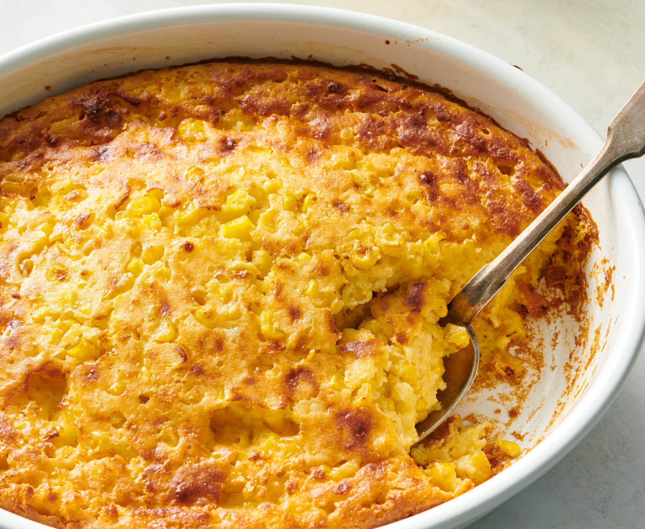 An oval baking dish full of corn pudding with corn kernels. There is a serving spoon in the dish.