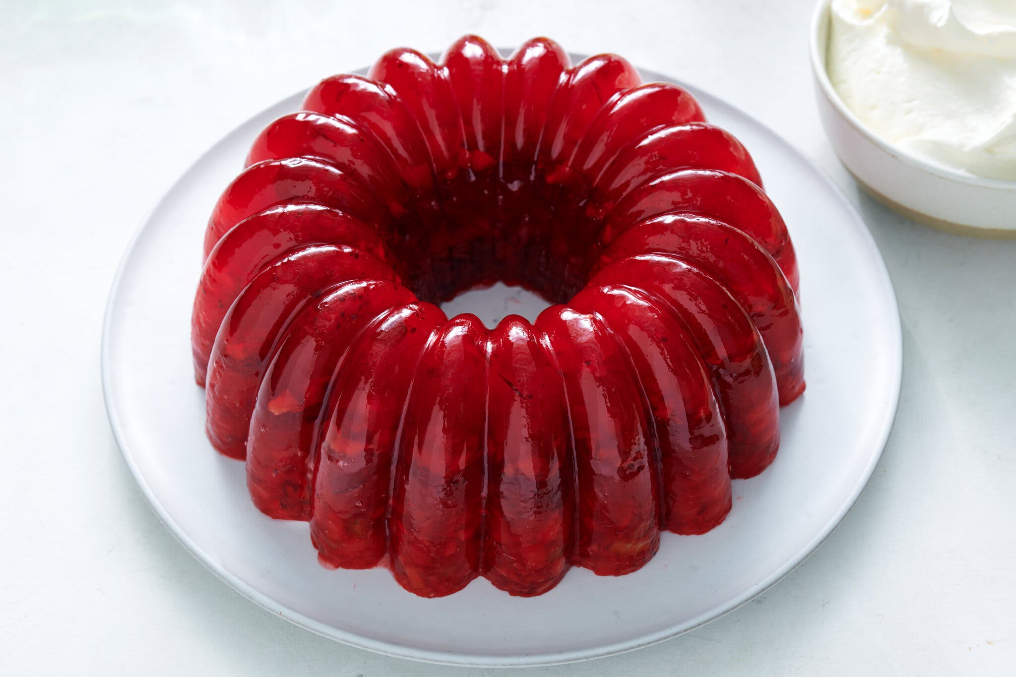 A shiny, red Bundt jelly sits on a white plate. There is a dish of whipped cream in the top right corner.