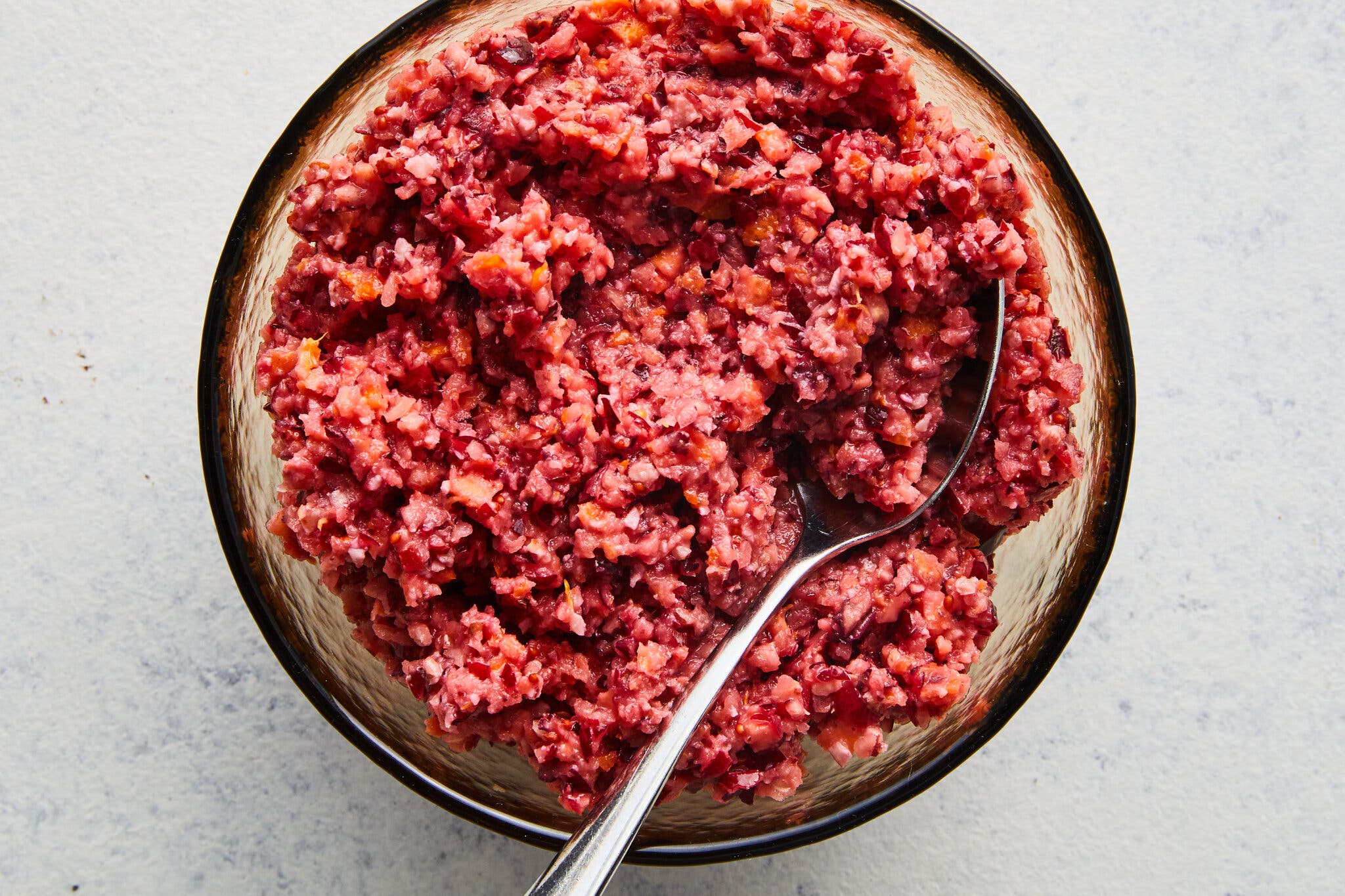 A clear glass bowl holds a red, coarsely chopped cranberry relish. A silver spoon rests in the mixture.