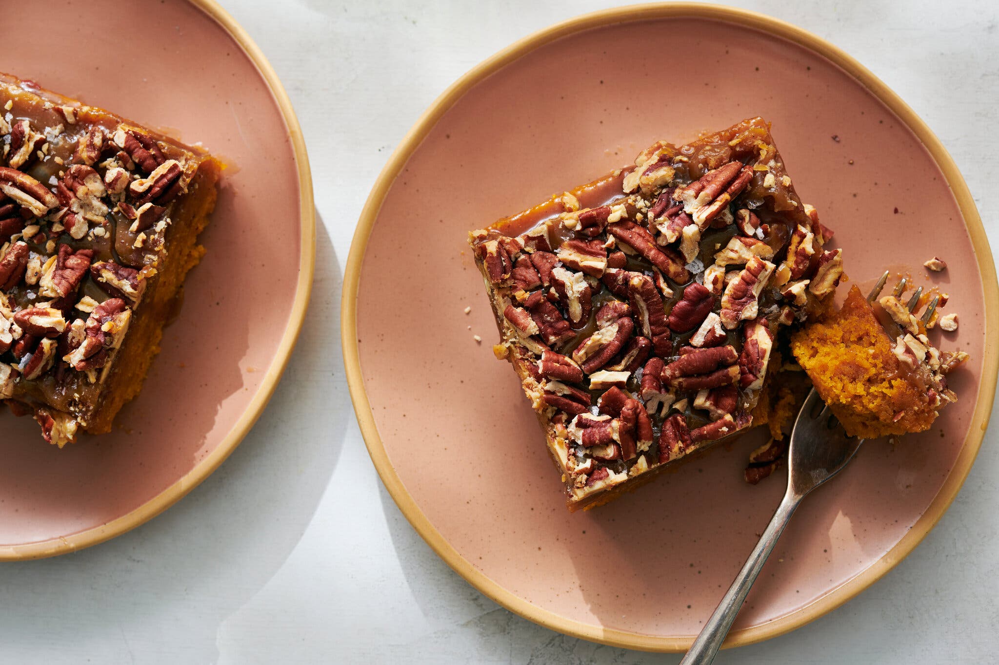 Two square slices of orange cake topped with caramel and pecans sit on pink plates. A fork removes a piece from one slice of cake.