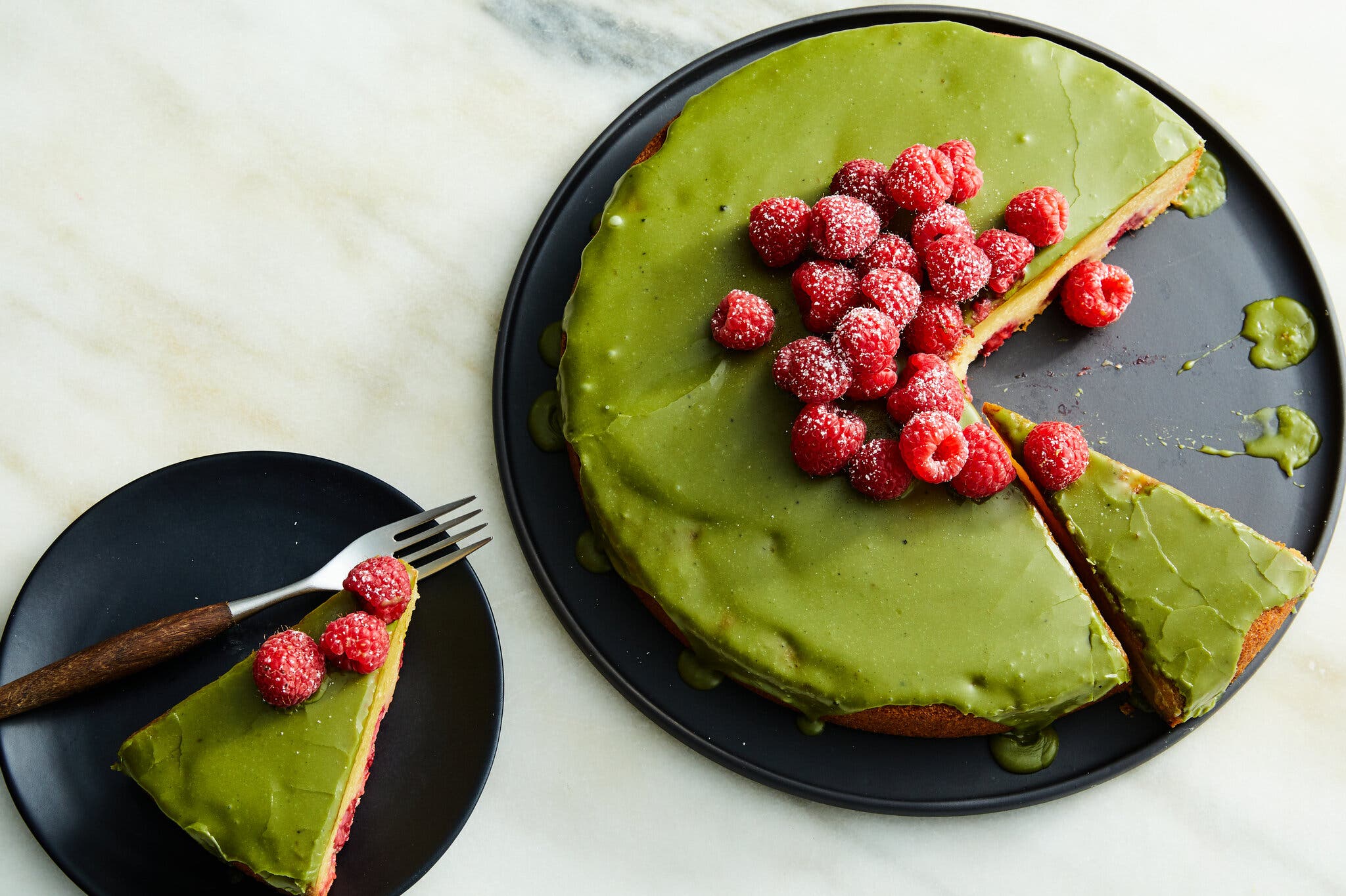 A round cake topped with green frosting and red raspberries sits on a black plate. Several slices have been cut, and one sits on a plate nearby.