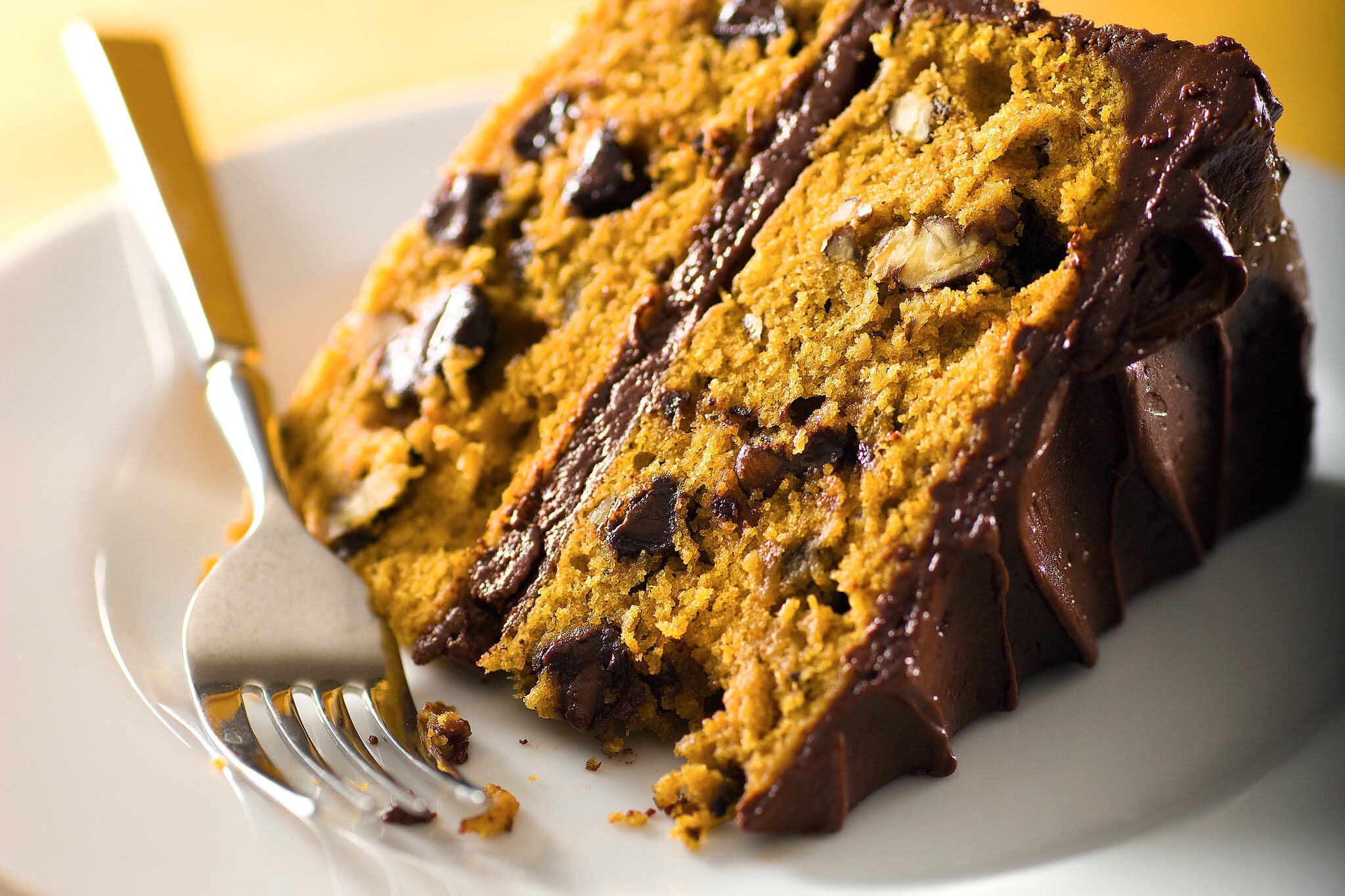 A slice of chocolate-frosted pumpkin cake studded with nuts and chocolate. A silver fork sits on the plate next to it.