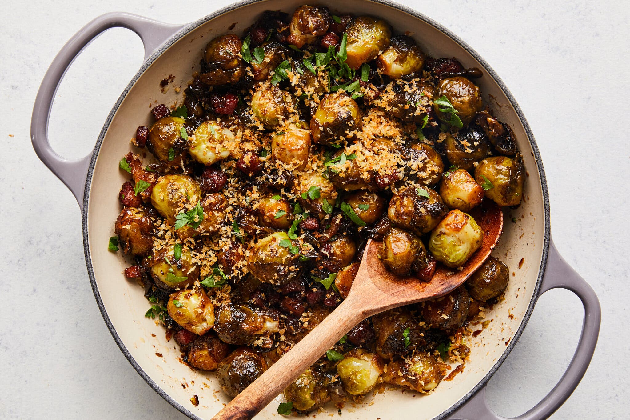Roasted brussels sprouts with pancetta, garnished with panko breadcrumbs and parsley, served in a light gray enamel pan with a wooden spoon.