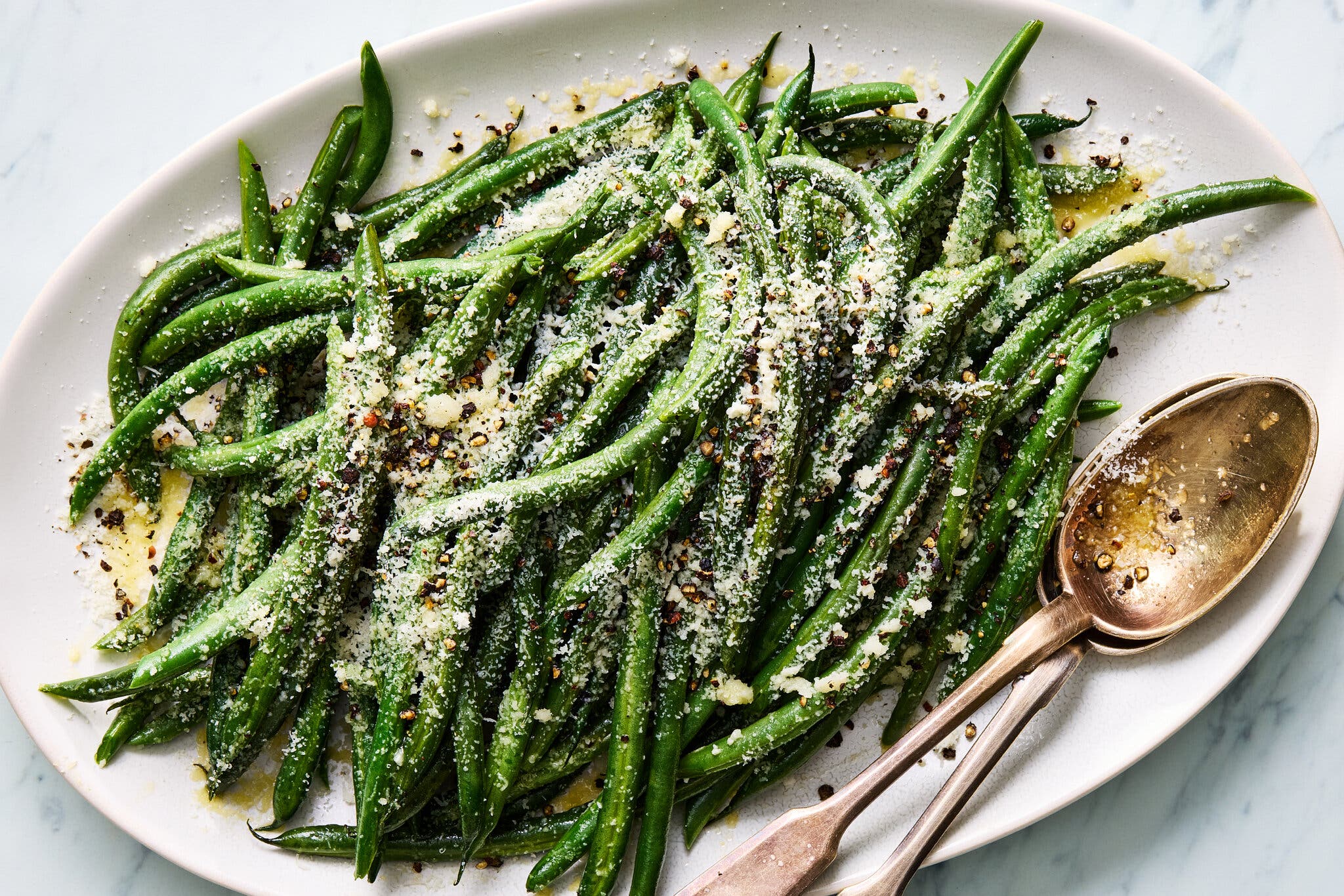 Bright green beans sprinkled with grated Parmesan and black pepper, served on a white oval platter next to a silver serving spoon.