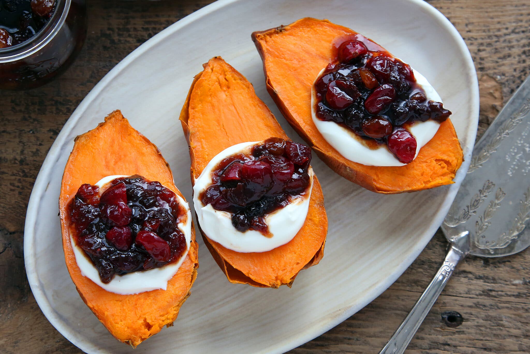 Three halved sweet potatoes, topped with cream and a cranberry chutney, on a white platter photographed from an angle above.
