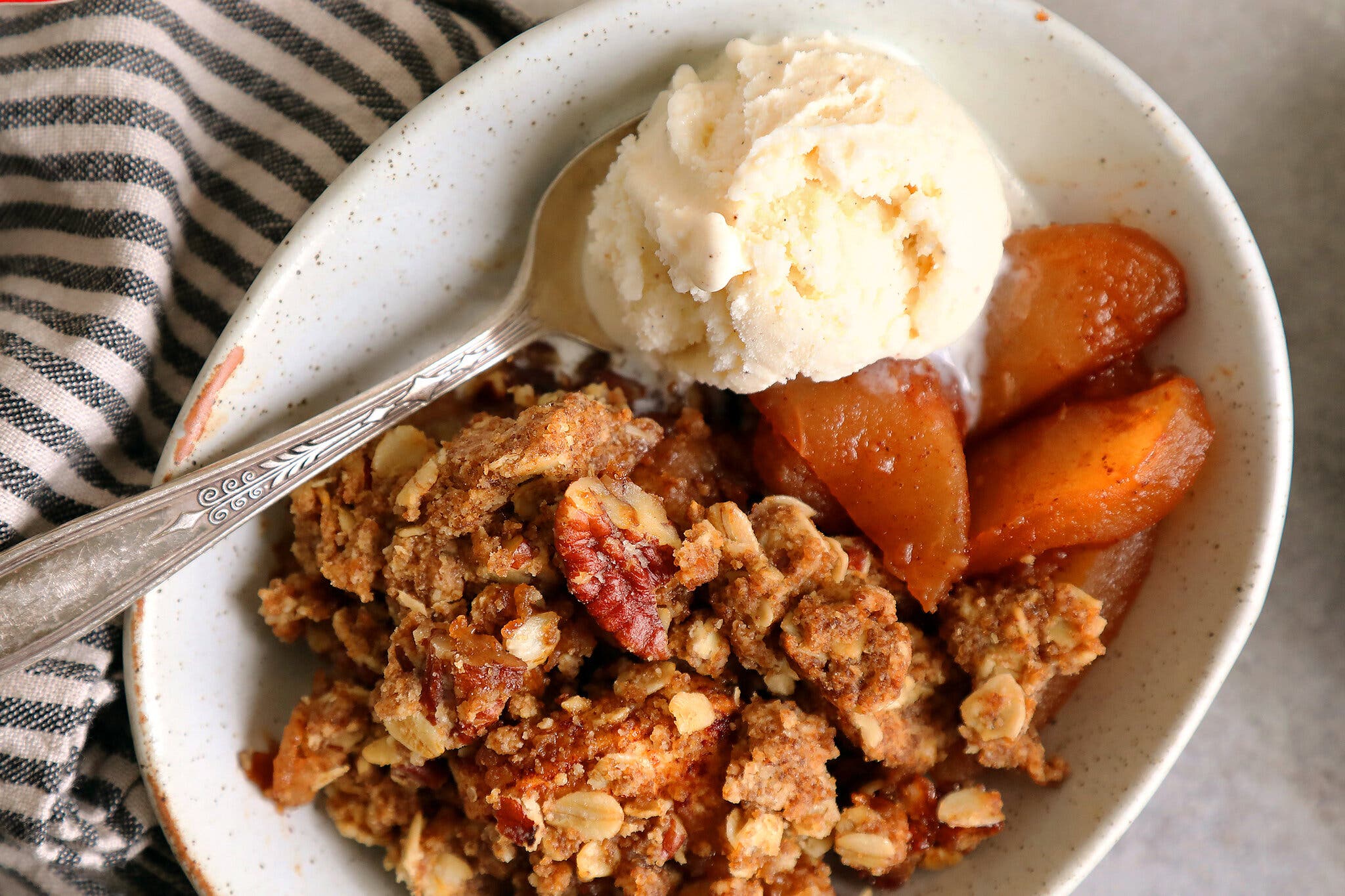 An overhead image of an oval bowl filled with apple crisp and a scoop of vanilla ice cream. A silver spoon is dipped into the ice cream. 