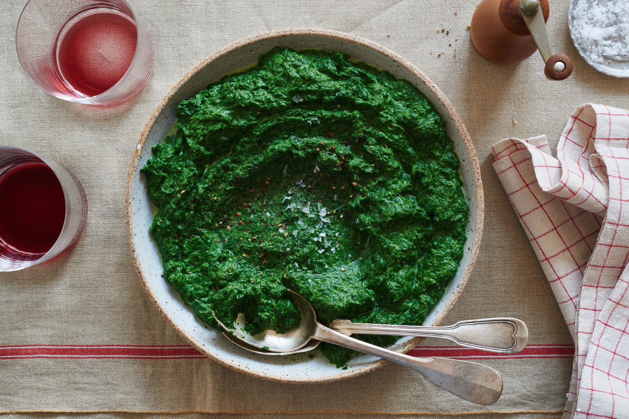 An overhead image of a white ceramic bowl on top of a light brown linen tablecloth with a red line. The bowl is filled with bright green creamed spinach sprinkled with flaky sea salt and ground black pepper. A silver spoon and fork are tucked into the spinach at the bottom of the bowl.