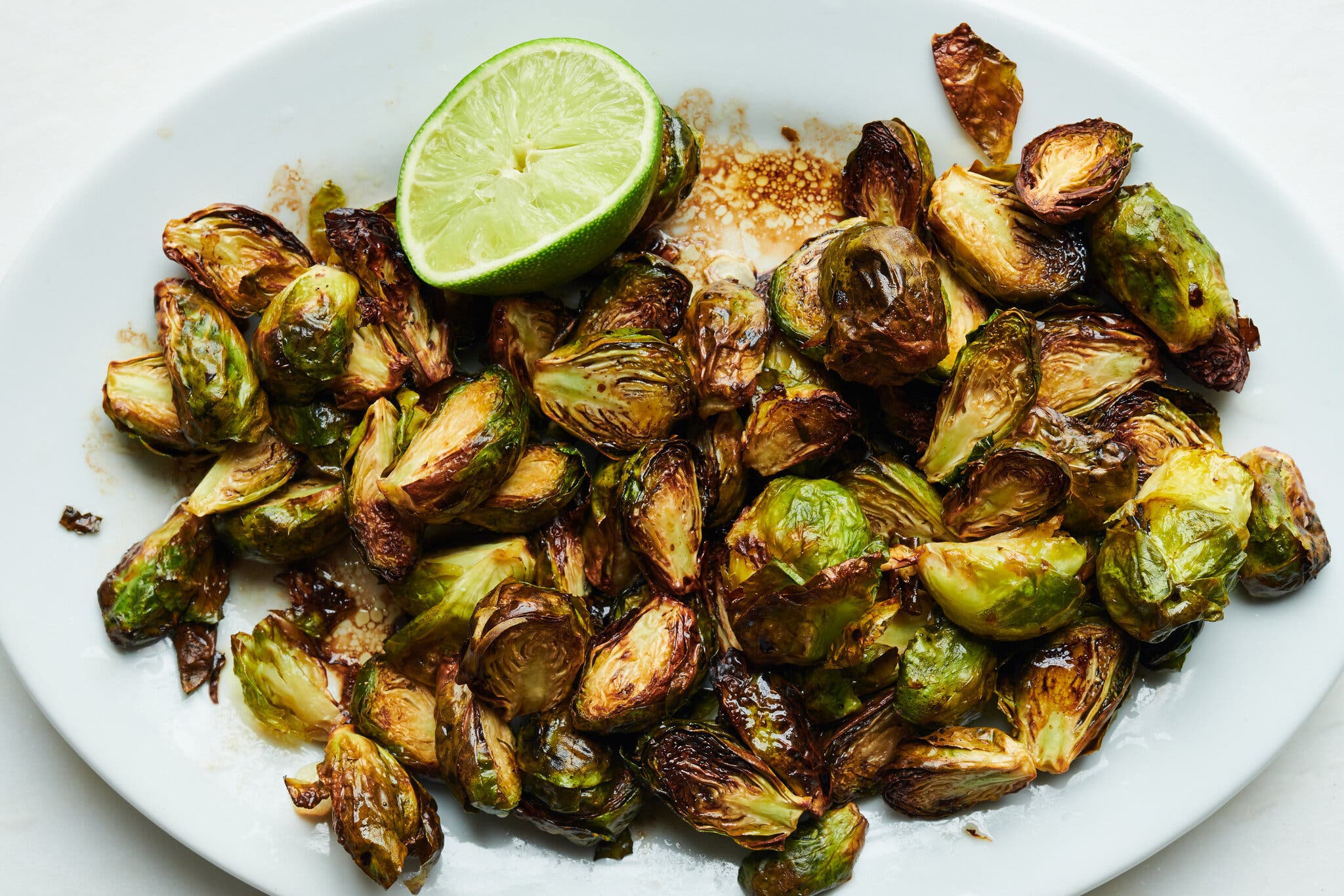 An overhead image of halved brussels sprouts with lightly charred edges on a white platter. A spent lime half sits to the top of the frame.