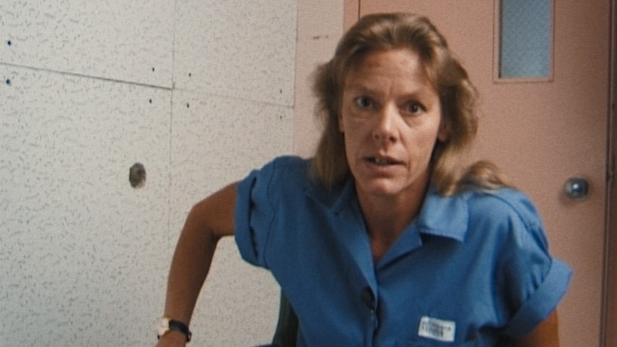Aileen Wuornos in a blue jumpsuit getting up from her seat.