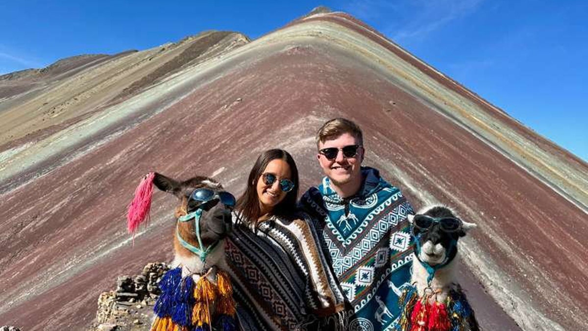 wo people wearing patterned ponchos pose with two llamas wearing sunglasses and colorful decorations in front of the multicolored slopes of Rainbow Mountain in Peru.