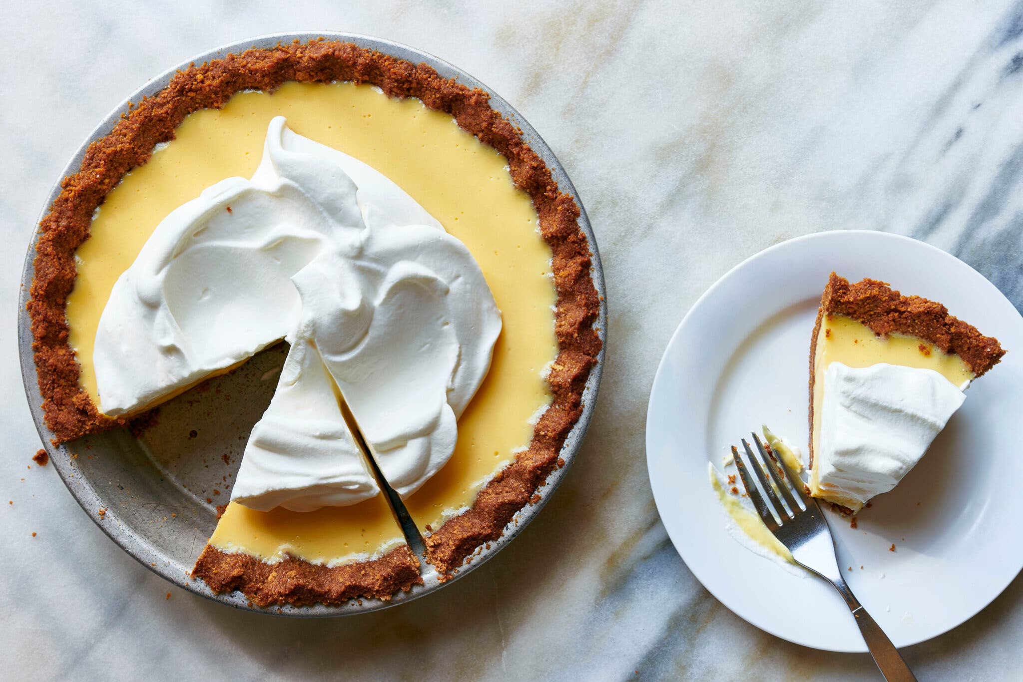An overhead image of a Key lime pie topped with whipped cream in a silver pie dish. A single slice is removed to a white plate with a fork on the right side of the image.