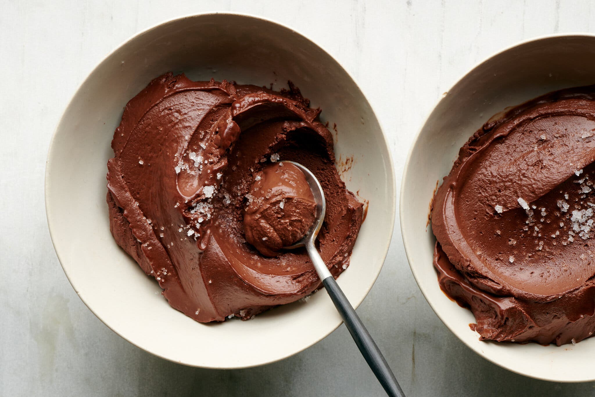 An overhead image of two white bowls filled with chocolate mousse sprinkled with flaky sea salt. A spoon is tucked into one of the bowls.