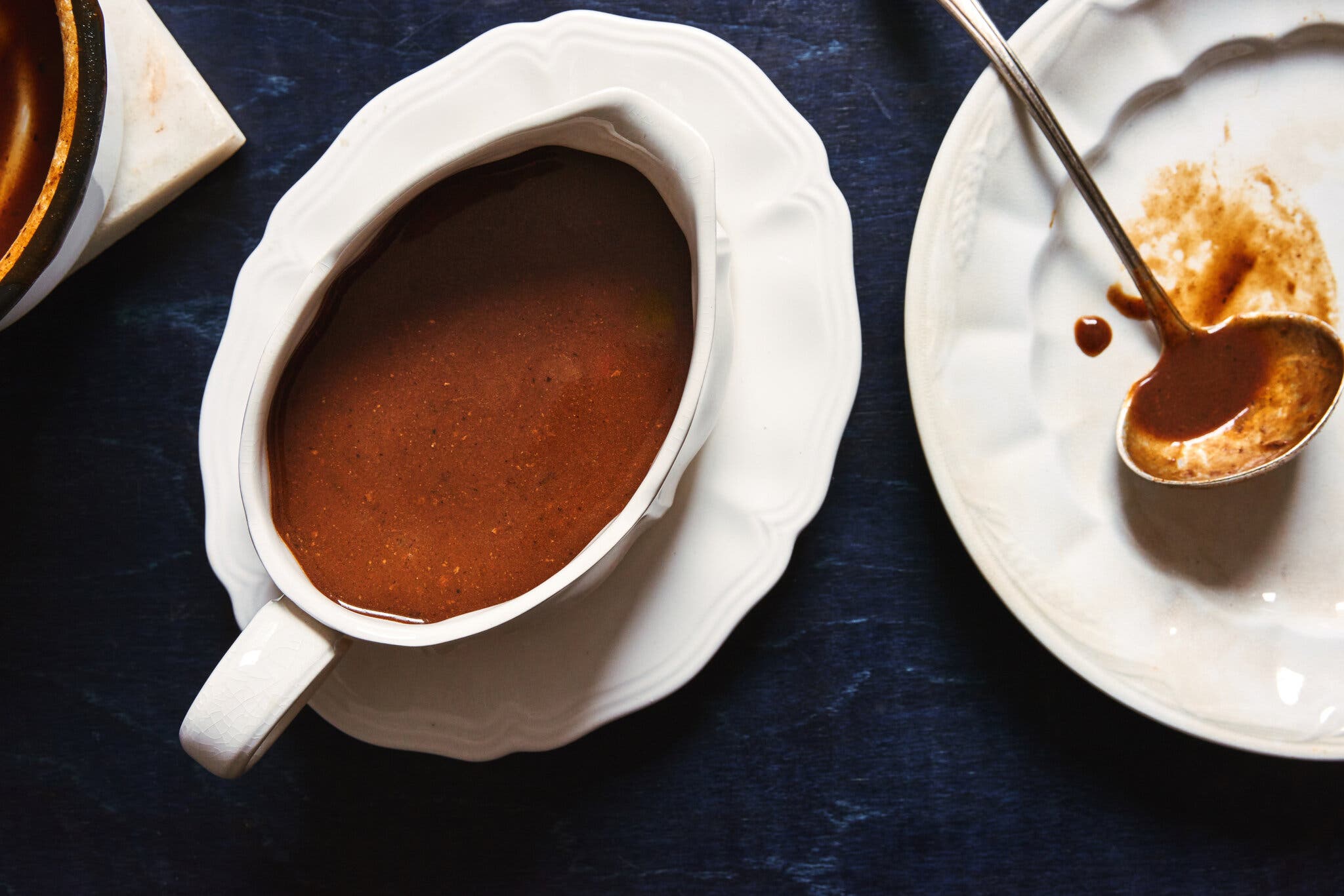 A white gravy boat sits on a plate against a dark blue surface. To the right is a plate holding a serving spoon that’s been coated in gravy.