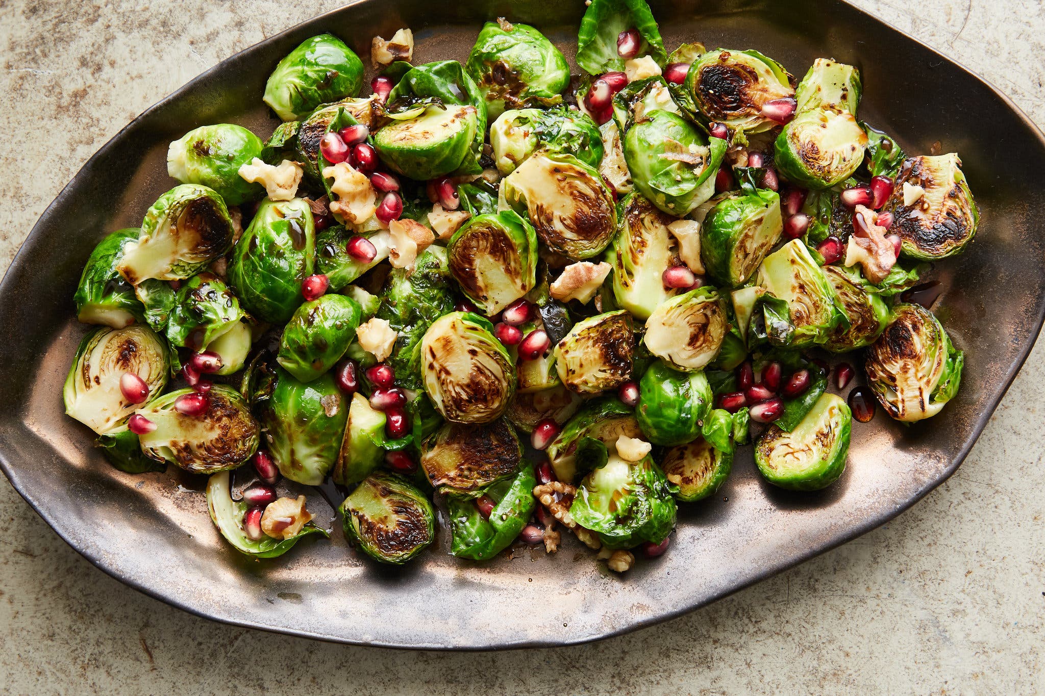 An overhead image of a dark gray oblong serving platter filled with seared brussels sprouts, walnuts and pomegranate seeds.