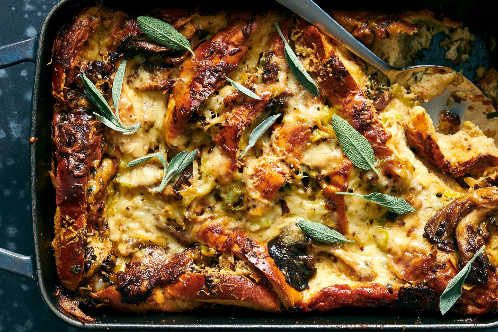 An overhead shot of a baking dish of bread pudding, studded with mushrooms and topped with melted cheese and sage leaves.