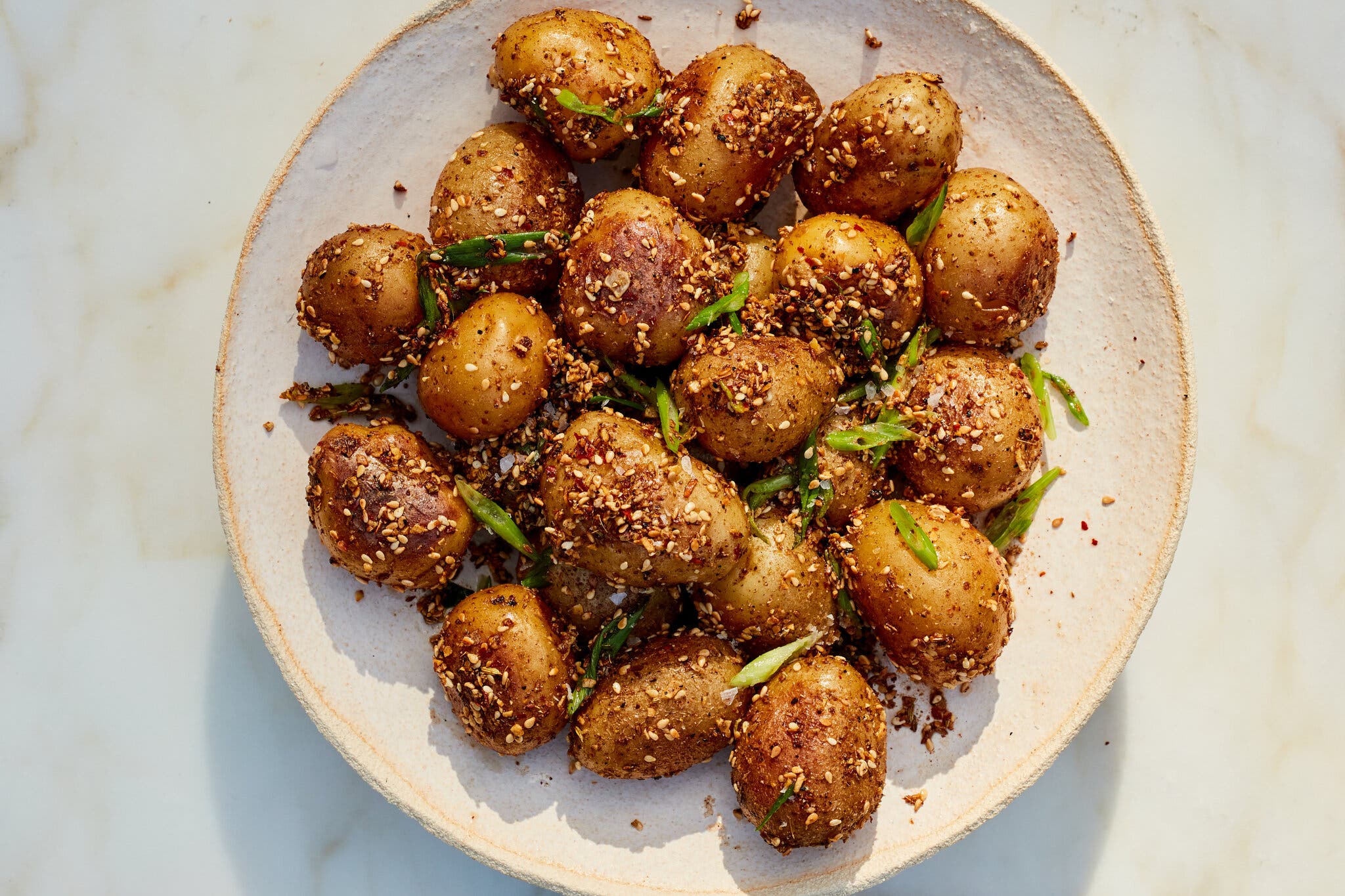 A light tan plate full of cooked new potatoes topped with sesame seeds and sliced shallots sits against a white marbled surface.