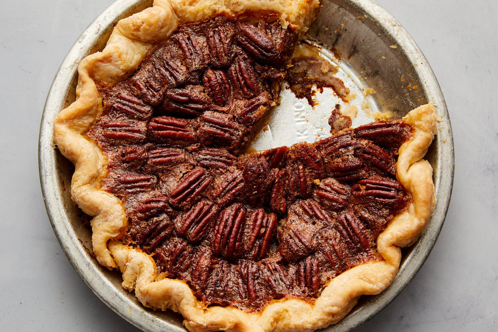 A pecan pie in a metal pie plate and shot overhead sits against a light gray countertop. One slice is missing.