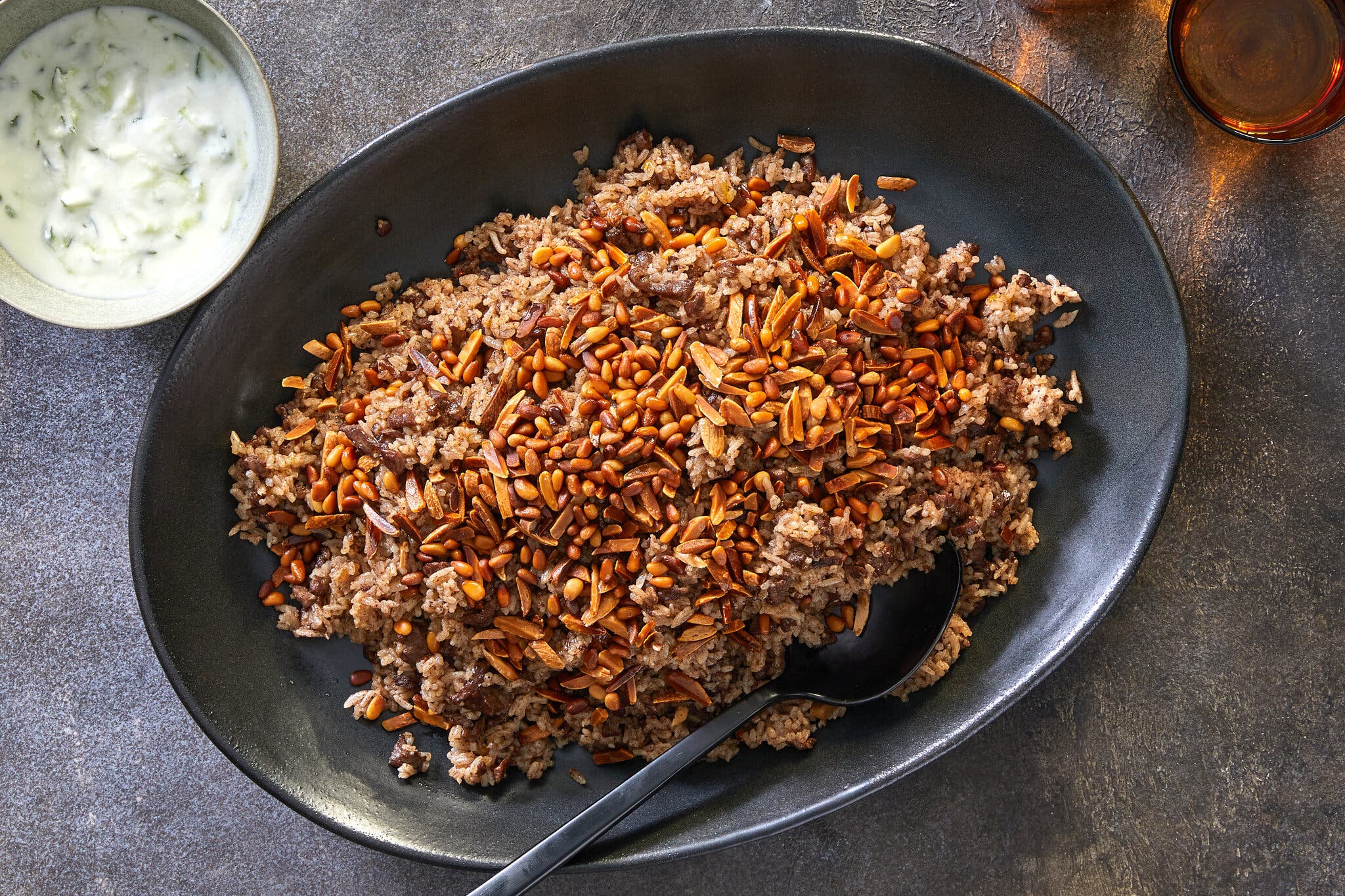 A large dark gray platter holding hashweh topped with a mix of toasted nuts sits against a gray background. To the top left is a bowl of yogurt sauce. To the top right there is an amber-colored glass.