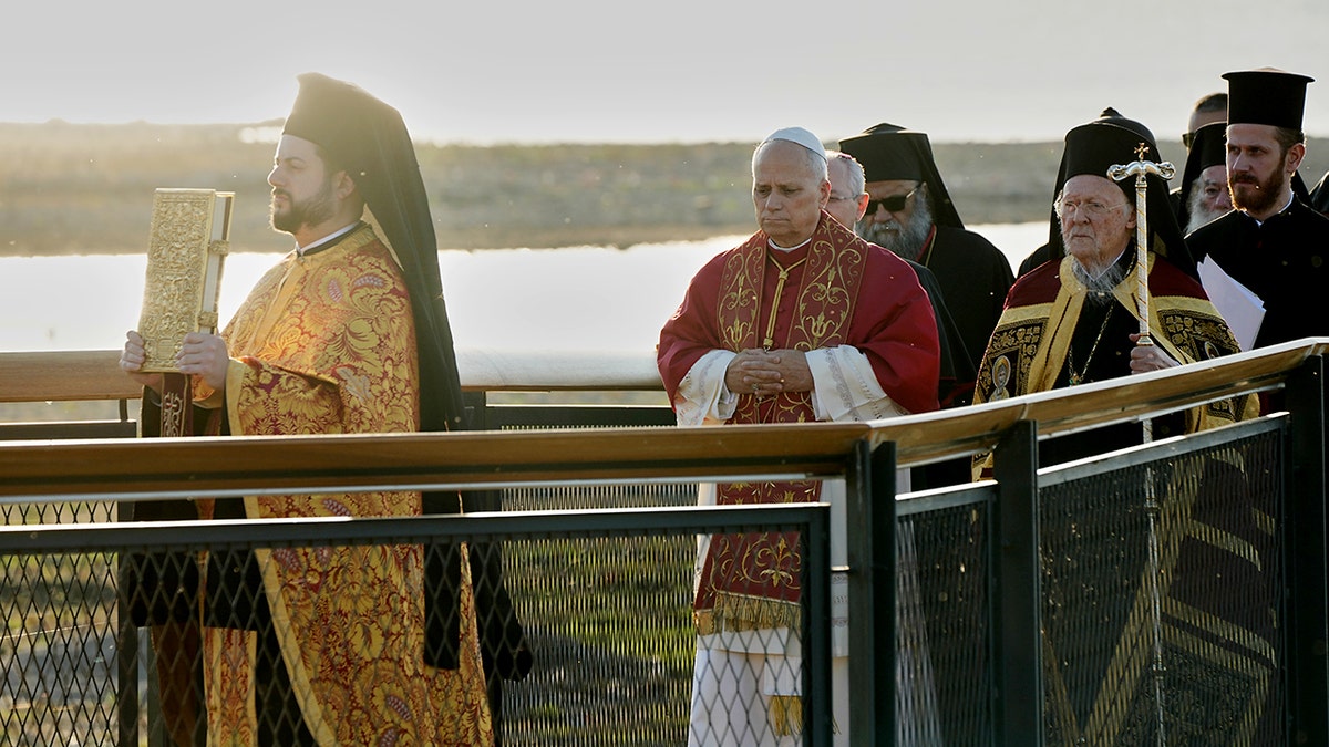 Clerics in ornate vestments walk in a religious procession along a waterfront platform.