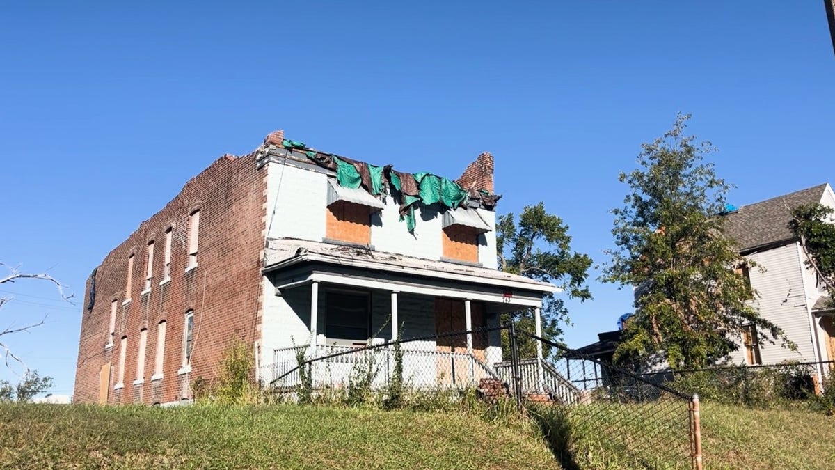 A two-story brick house in St. Louis stands with boarded windows and a torn tarp over its damaged roof months after the tornado.