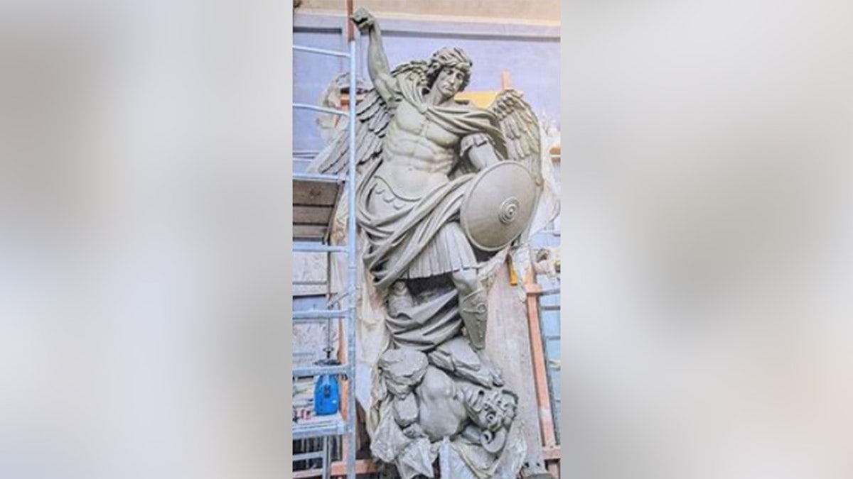 A large bronze sculpture of Saint Michael with wings extended, raising a spear and holding a shield while standing on a fallen figure, surrounded by scaffolding.