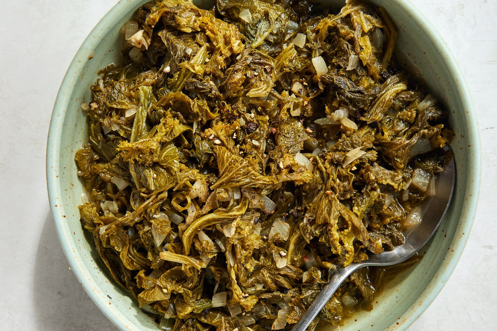 A light green bowl filled with cooked-down mustard greens sits against a light surface. Inside the bowl there is a spoon.
