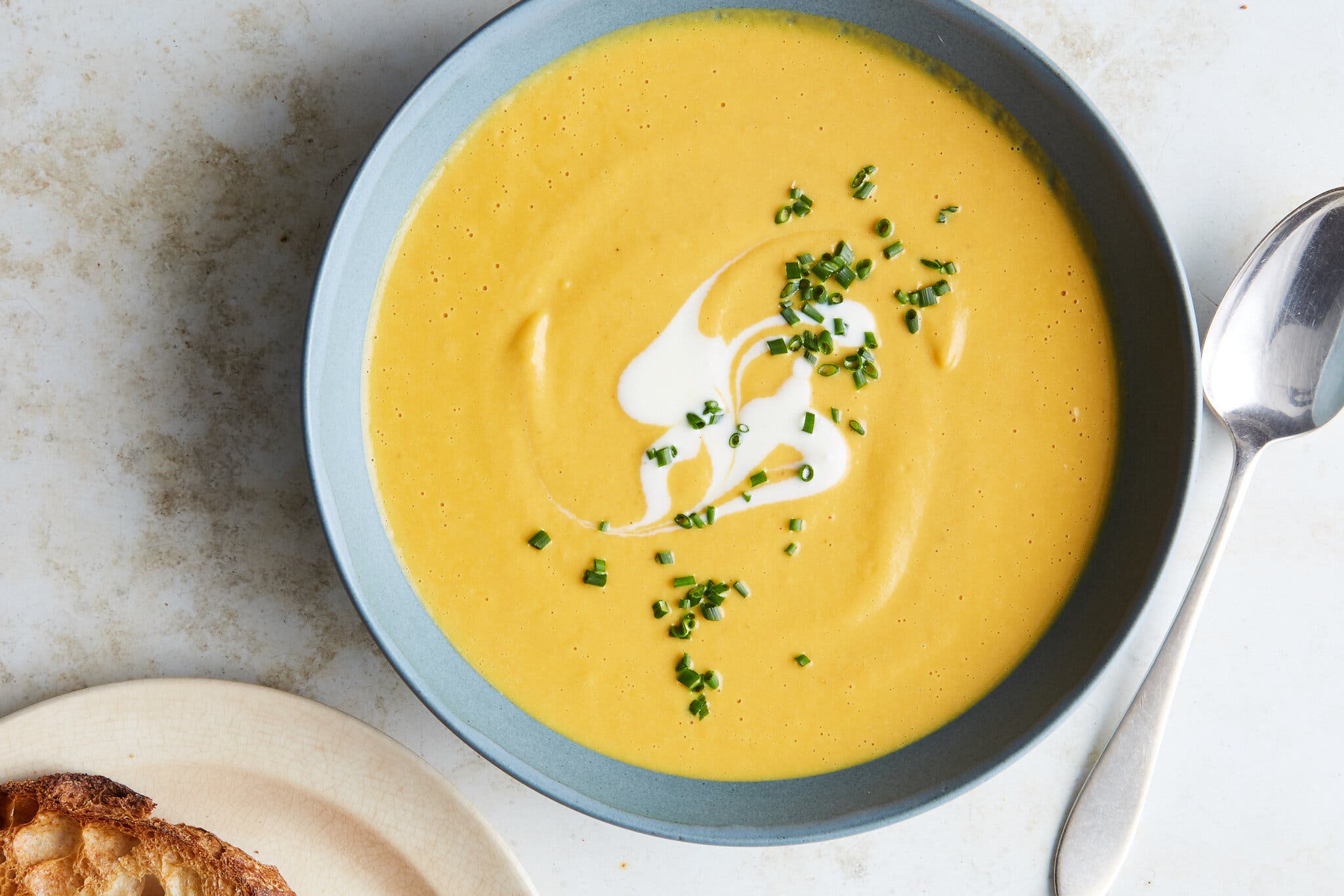 A bowl of bright yellow-orange soup topped with yogurt and chives sits on a light gray background. To the right is a spoon.