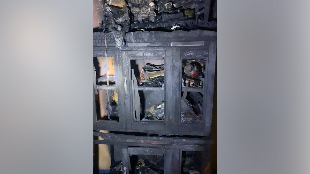 Burned wooden cabinet severely charred in a house fire, with melted objects, exposed wiring, and soot-covered shelves.