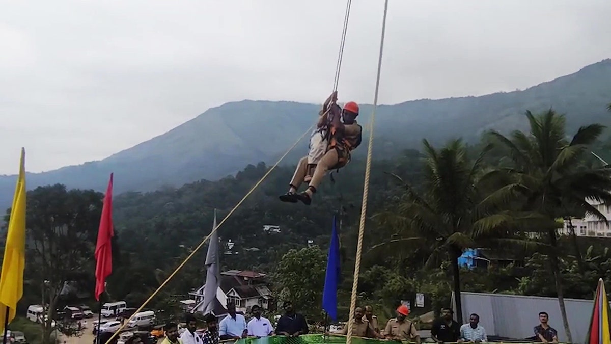 Firefighter rescues a family from sky-dining attraction by climbing a rope, while a crowd watches.
